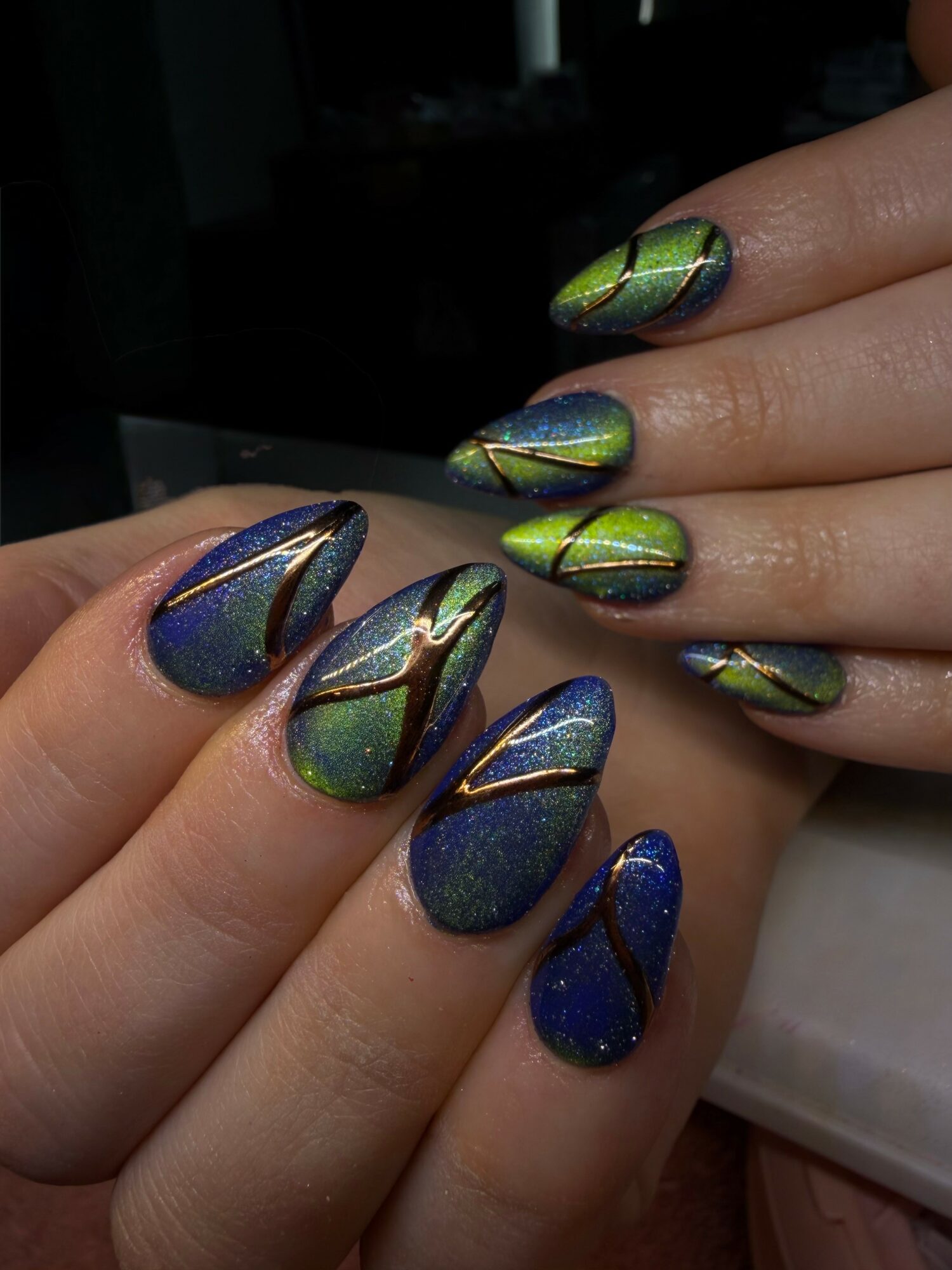 Close-up of hands with colorful, metallic, geometric-patterned nail art on fingernails, with a dark background.
