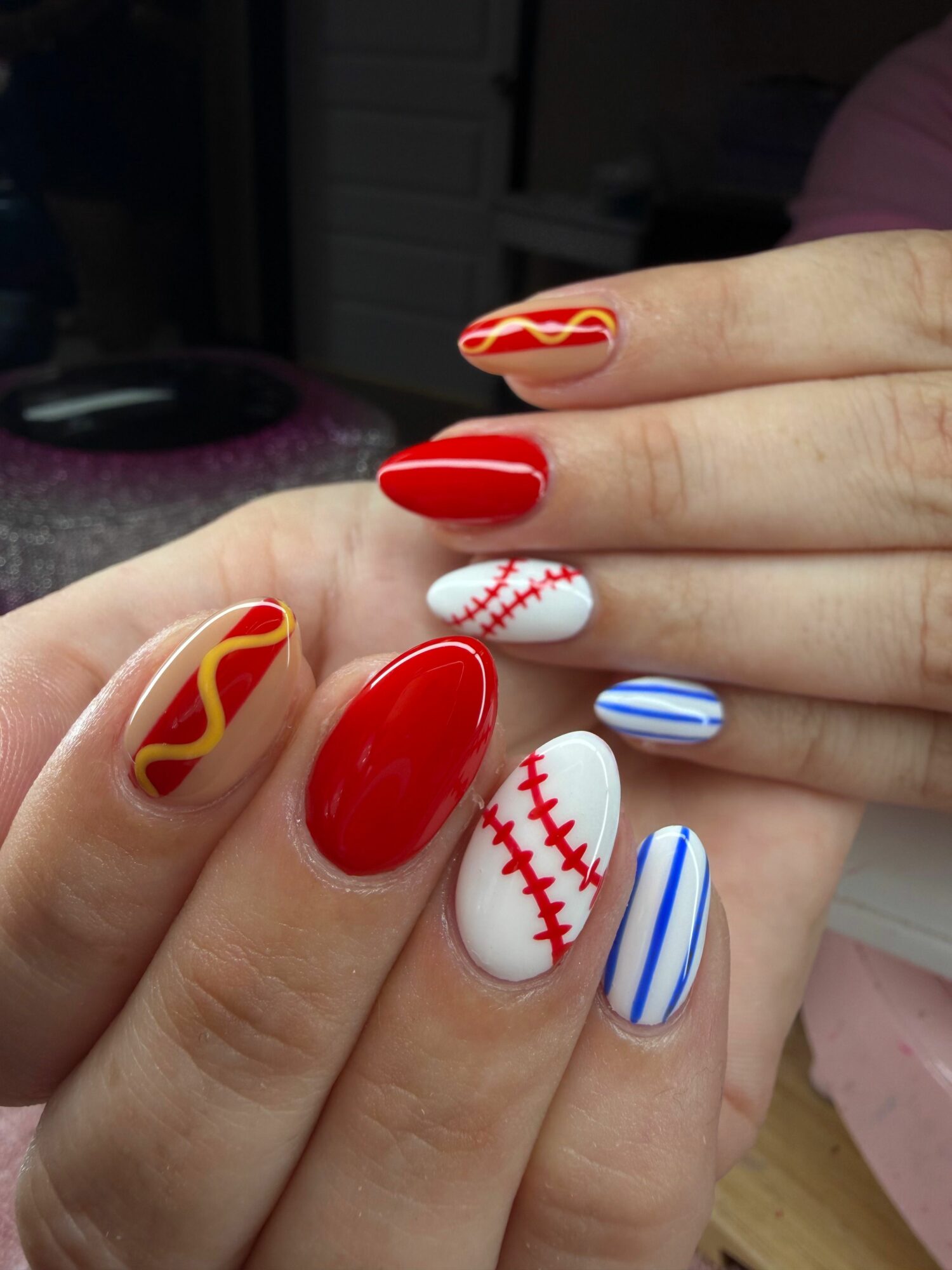 Hands with colorful baseball-themed nail art, including red, white, and blue designs, and a baseball pattern on one nail.