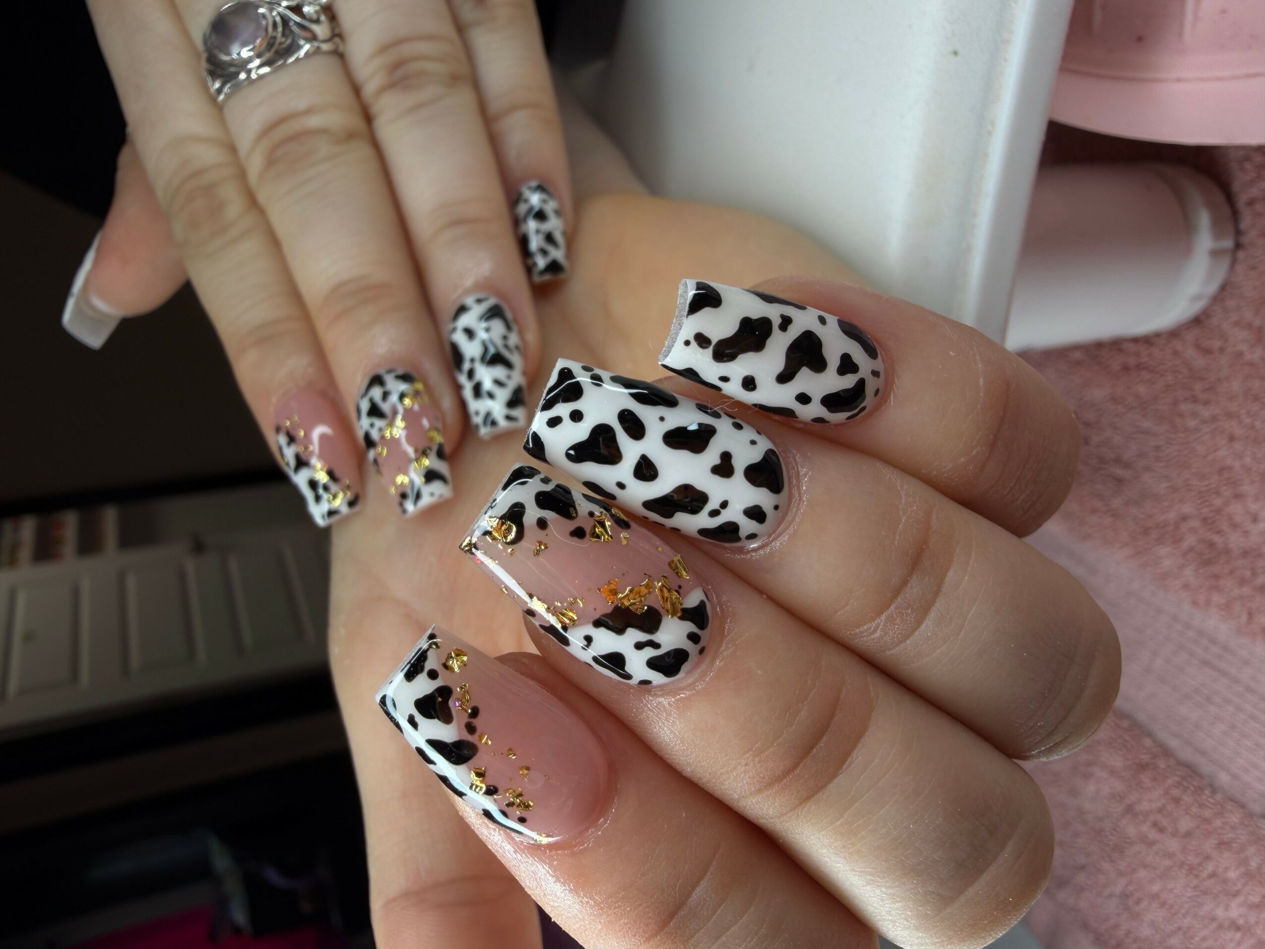 Hand with cow print and floral decorated nails, rings, and a pink towel in background.