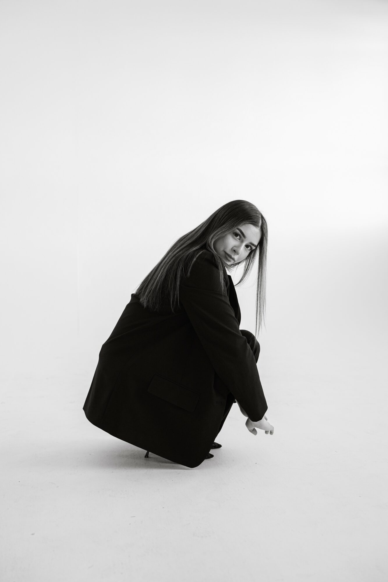 Person crouching on the floor, looking at the camera, with long hair and dark clothing, against a plain background.