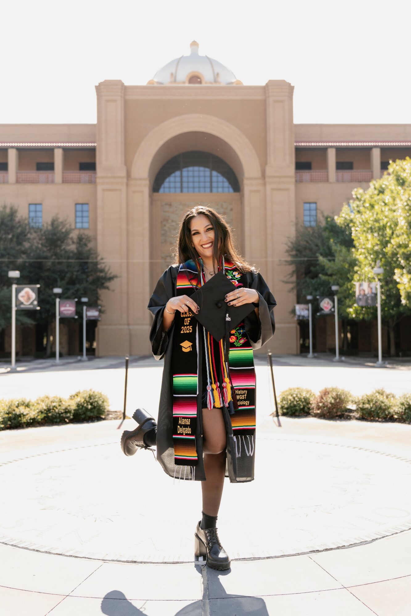 Graduate in cap and gown holding diploma in front of university building with trees and clear sky