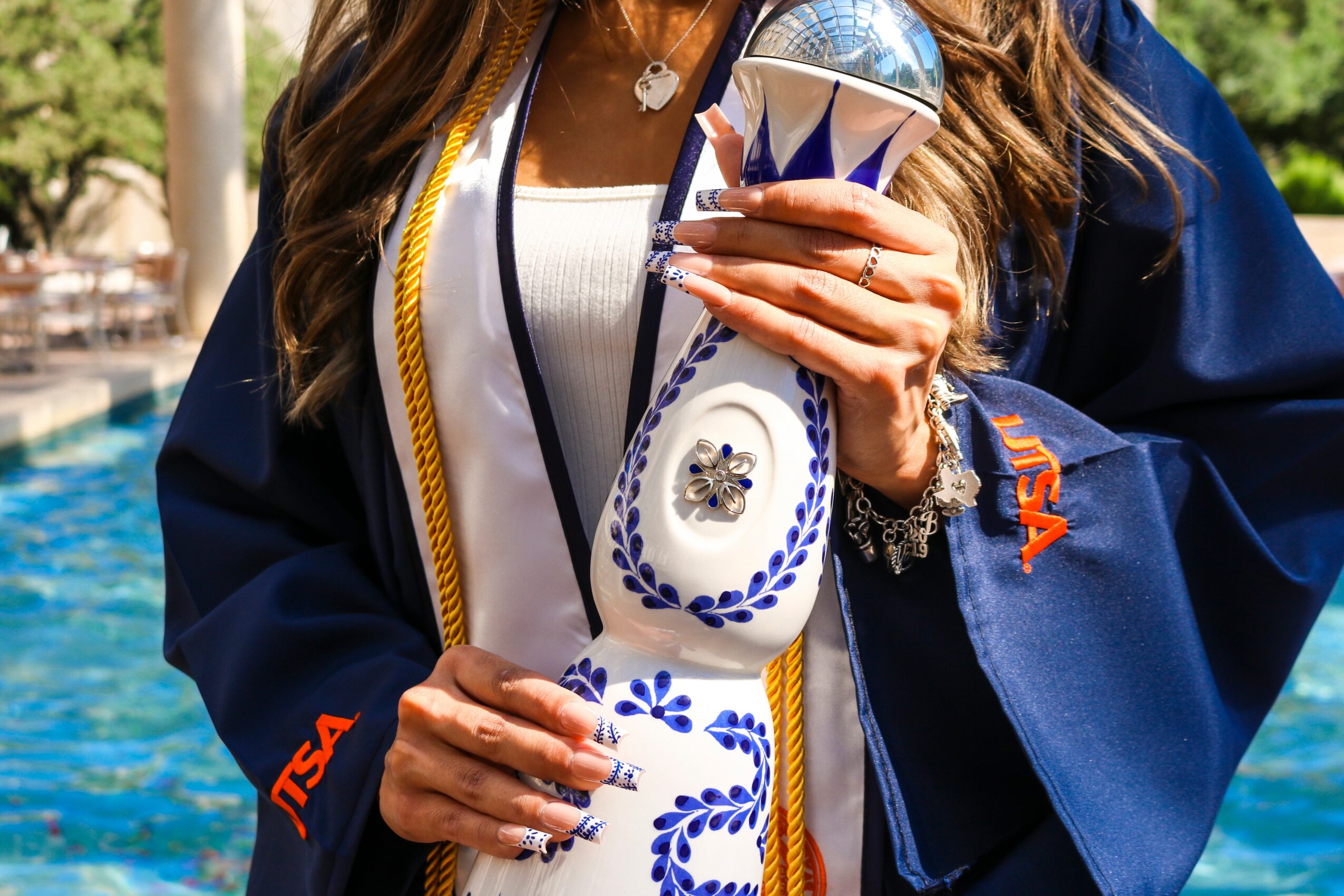 Person in graduation gown holding a trophy with a blue and white design, outdoors with trees and water in background.