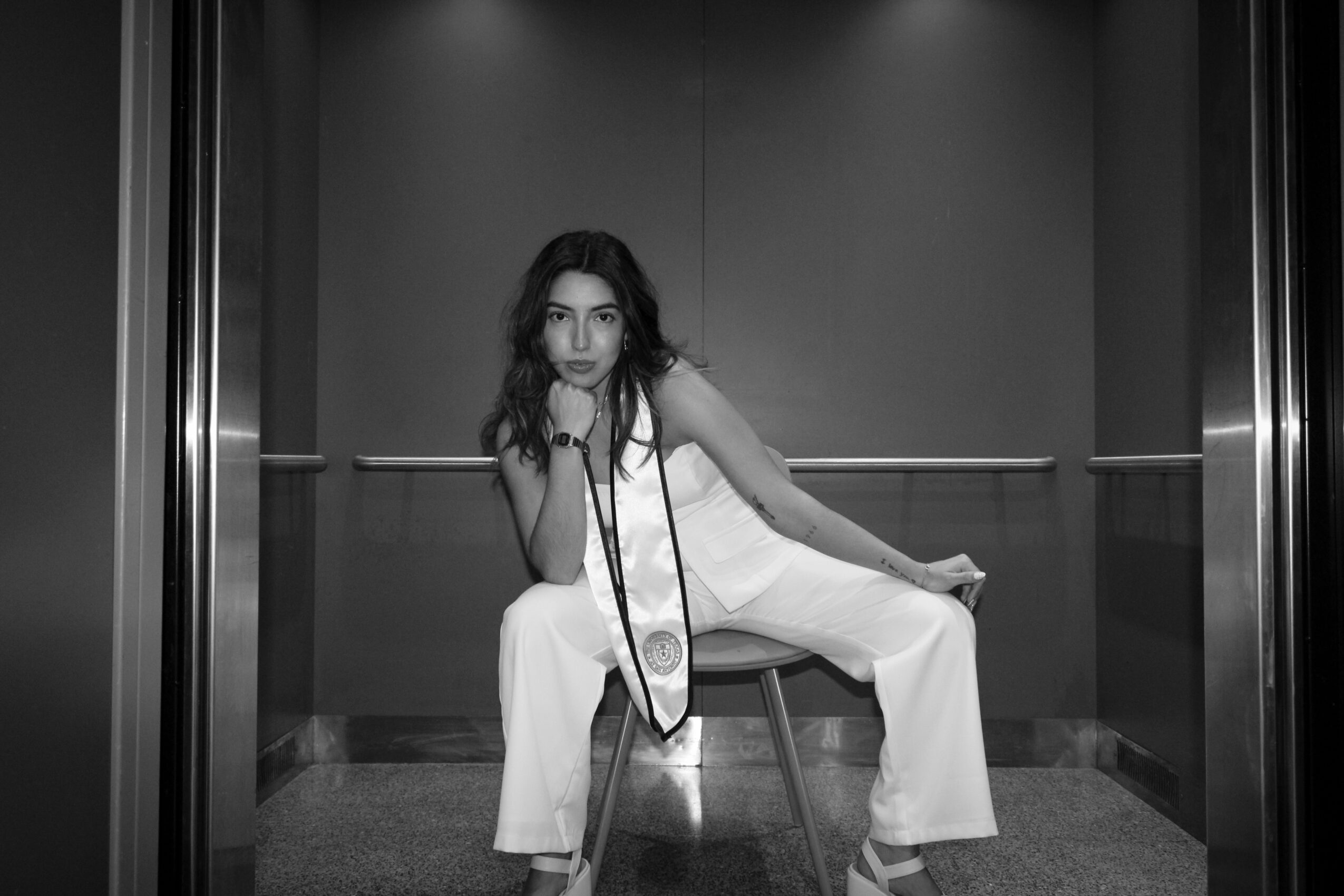Woman in white jumpsuit sitting on a chair in an elevator, holding a camera, with a neutral background.