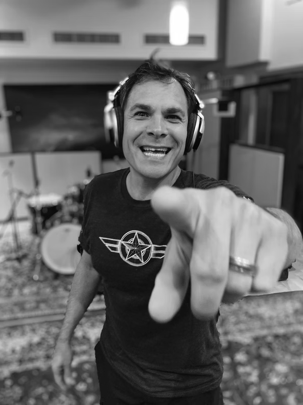 Man wearing headphones smiling and pointing at the camera in a recording studio, with musical instruments in the background.