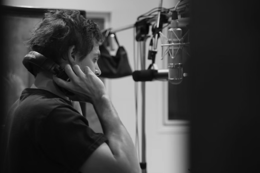 Person with short hair speaking into a microphone in a recording studio, holding headphones to one ear.