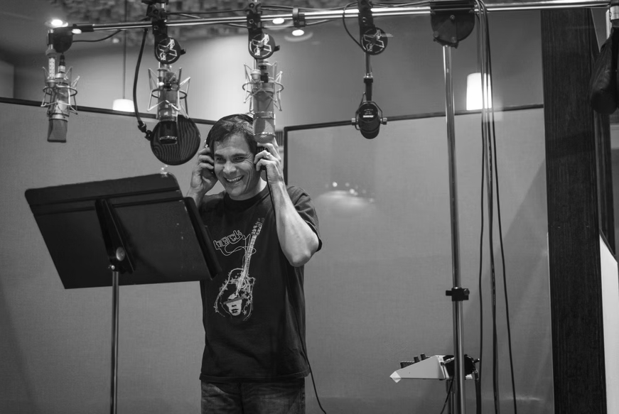 Person in a recording studio wearing headphones, smiling, standing in front of a microphone and music stand.
