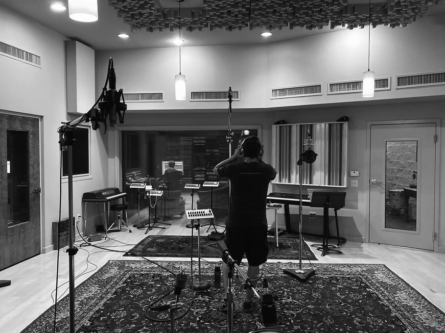 Person standing in a recording studio with microphones, music stands, and equipment, facing away from the camera.