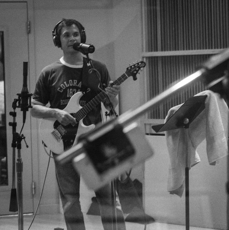 Person with headphones singing into a microphone and playing an electric guitar in a recording studio.