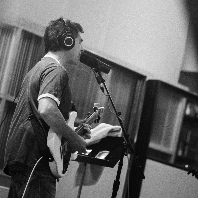 Person with headphones singing into a microphone while playing an electric guitar in a recording studio.