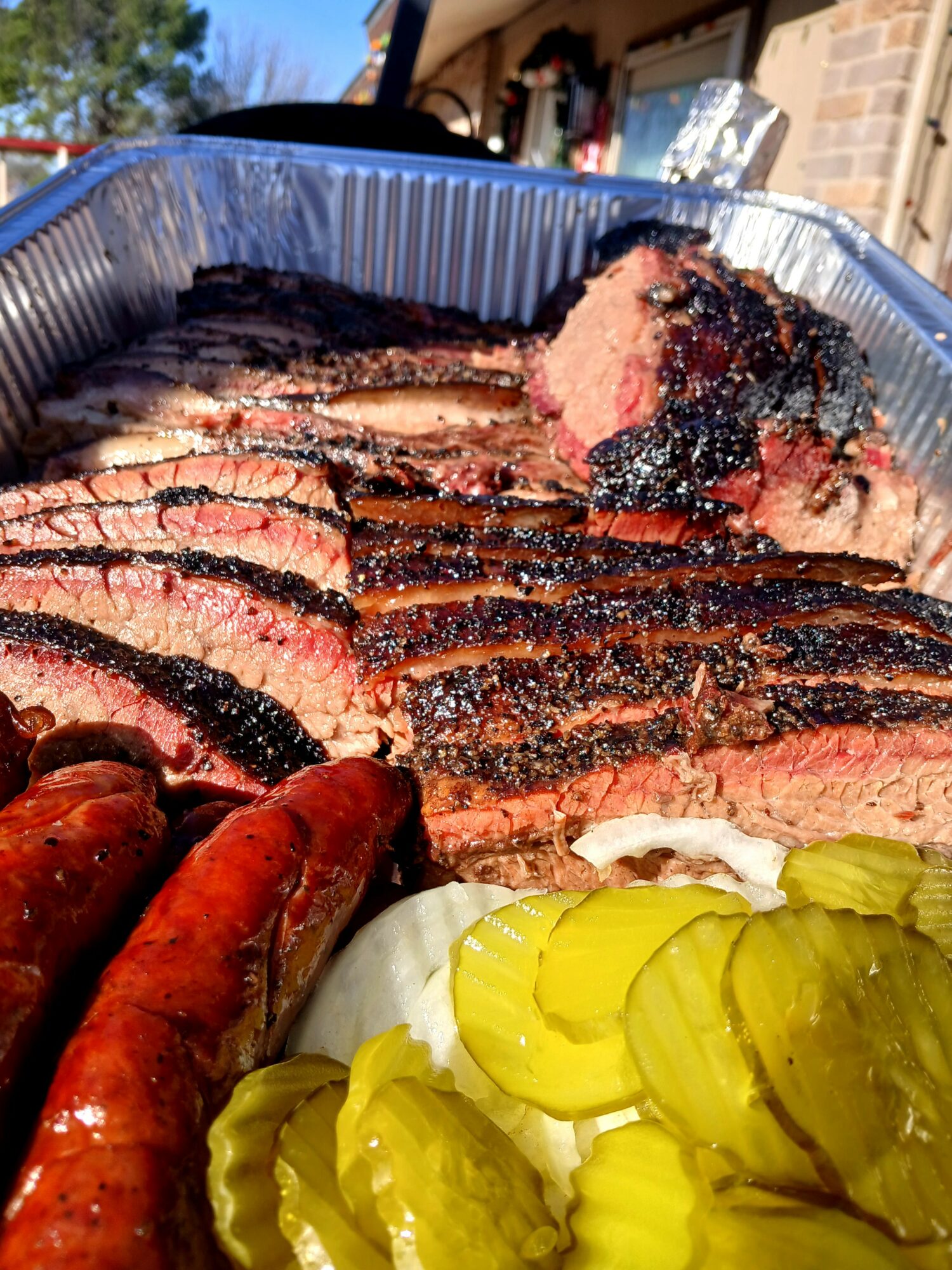 Sliced grilled steak with char marks, pickles, and sausages in a foil tray outdoors.