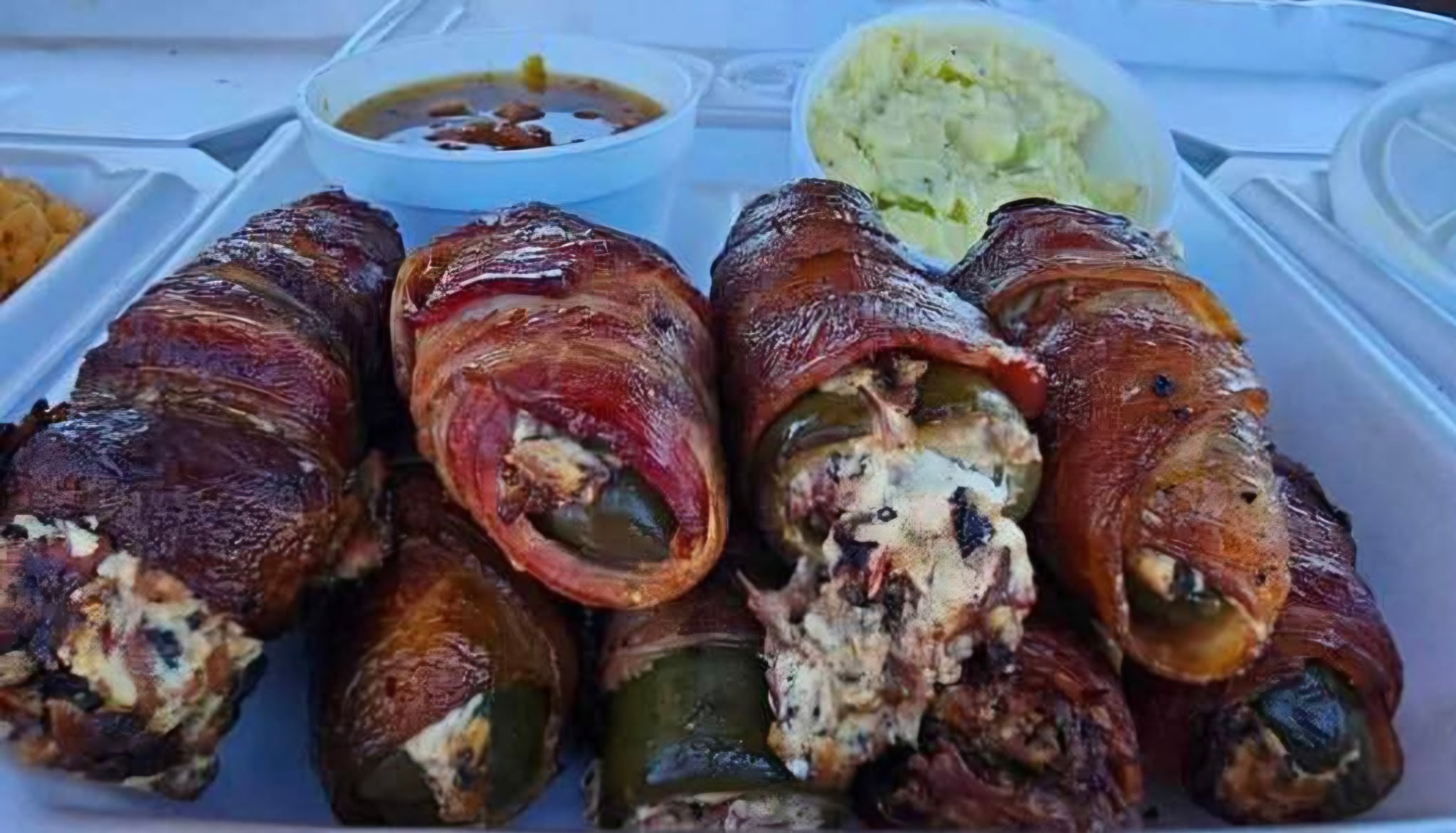 Six bacon-wrapped stuffed jalapeños on a tray with dipping sauces and shredded lettuce in the background.