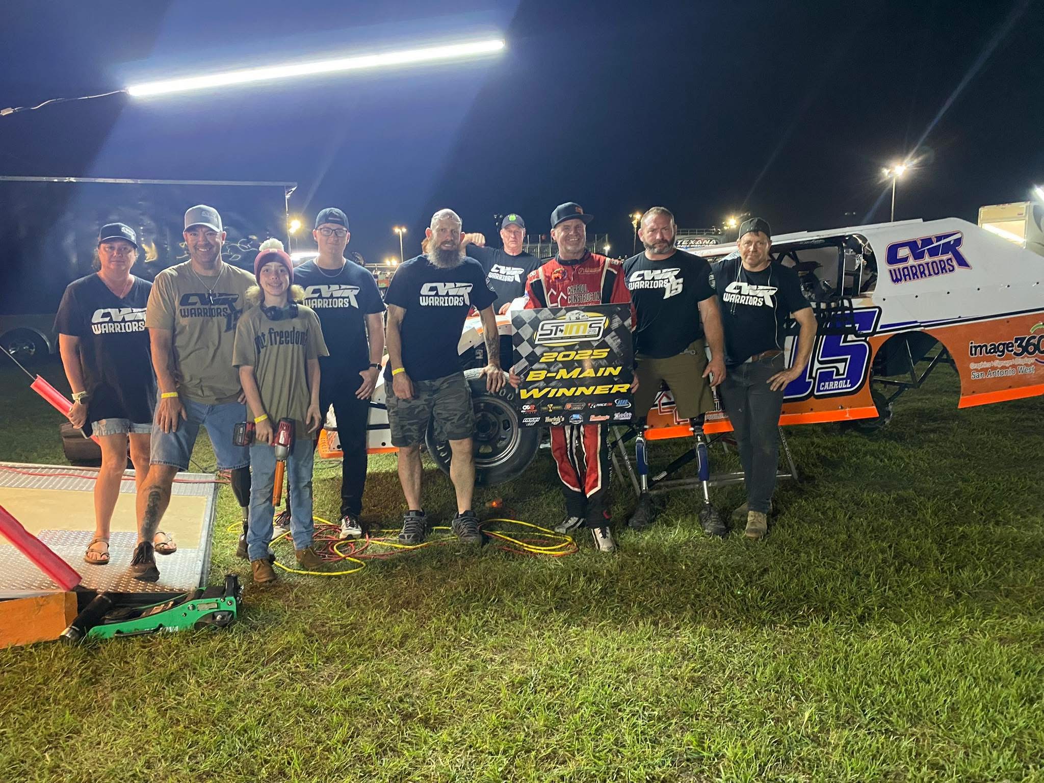 Group of people standing on grass near a race car at night, with bright lights overhead.