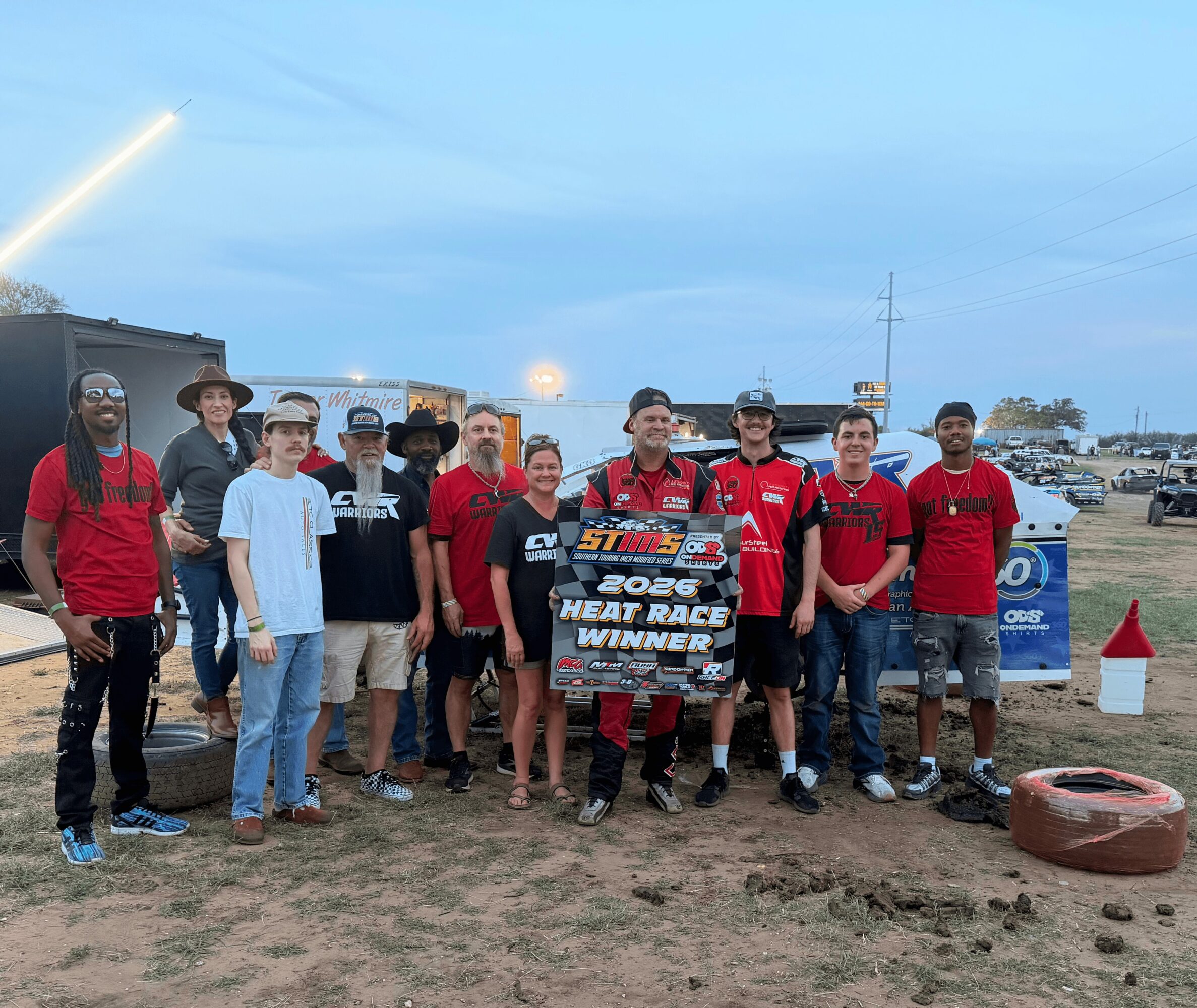 Group of people standing outdoors, holding a sign that reads '2003 Heat Race Winners,' with a race track and cars in the background.