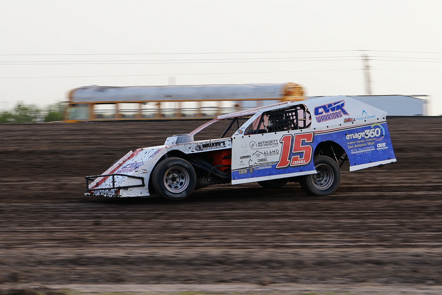 Race car with the number 15 speeds on a dirt track, with a train in the background.