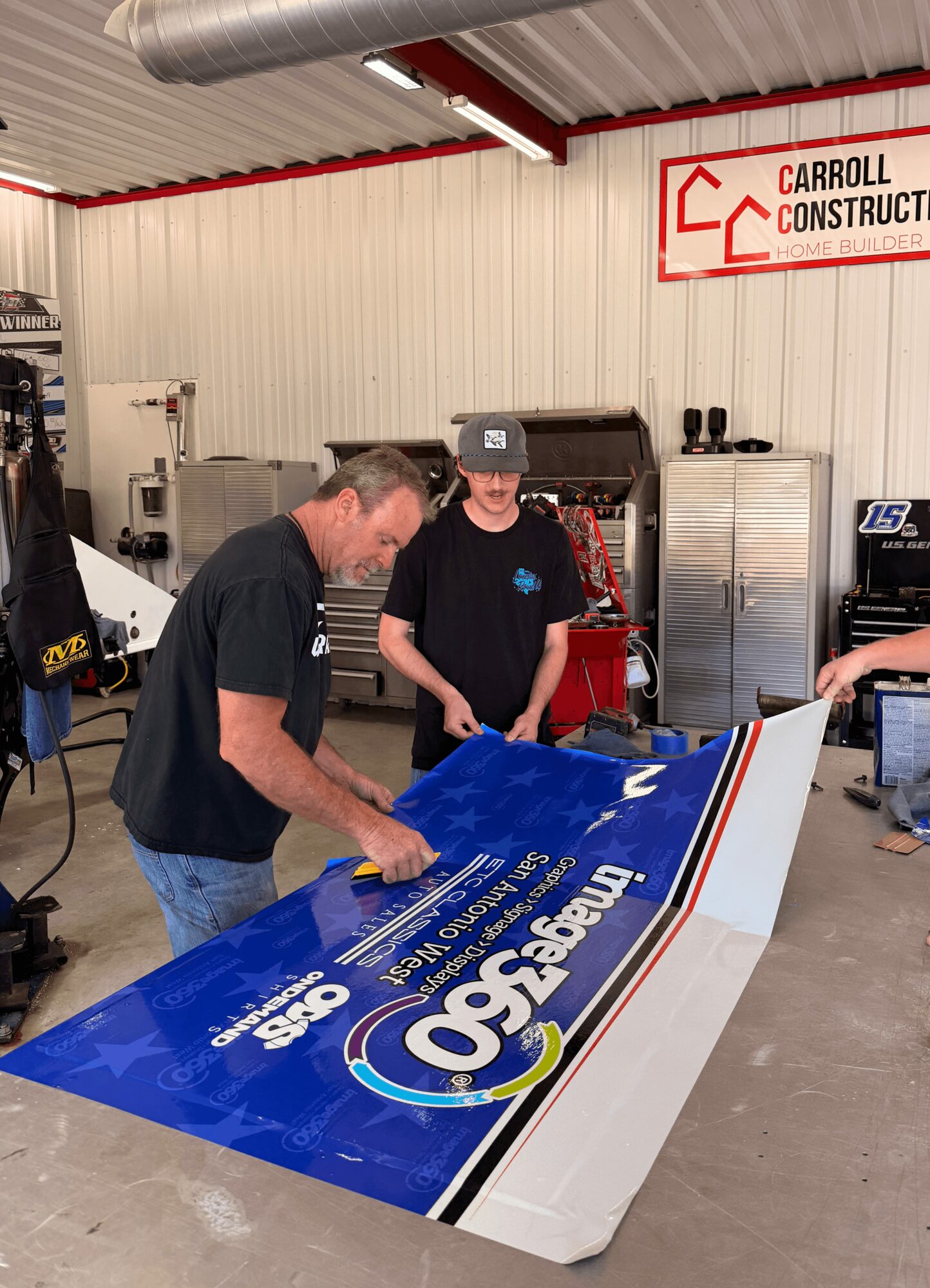 Two men examine a large blue and white sign in a workshop with tools and equipment, including a metal cabinet and a red sign reading 'Carroll Construct'.