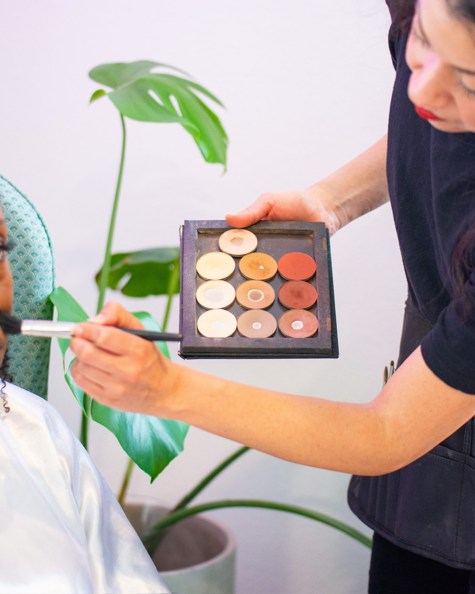 Makeup artist applying makeup to a person's face, holding a palette with various shades, with a plant in the background.