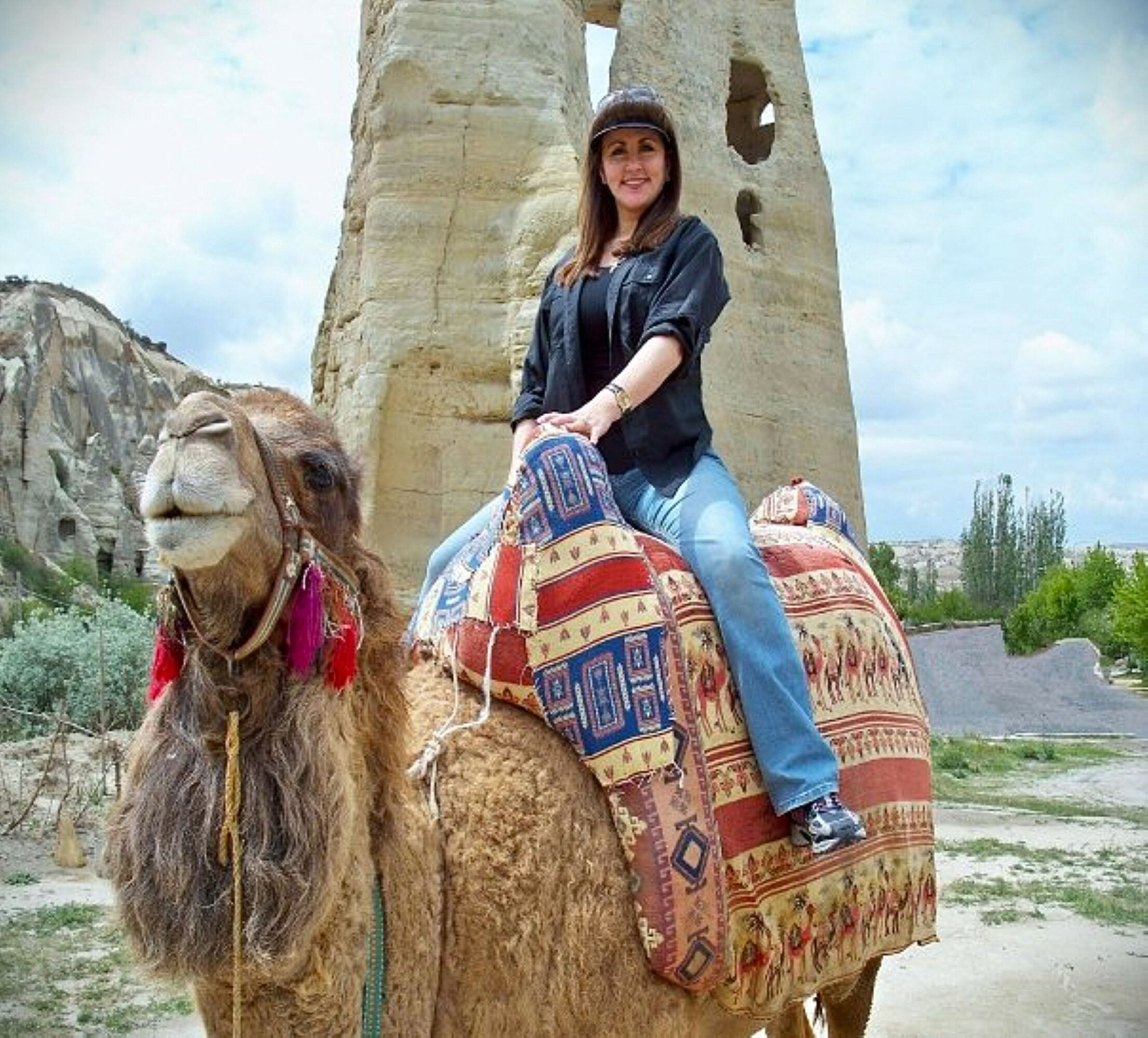 Woman riding a camel with a patterned saddle in a desert landscape, rock formations in the background, smiling, wearing a hat and casual clothes.