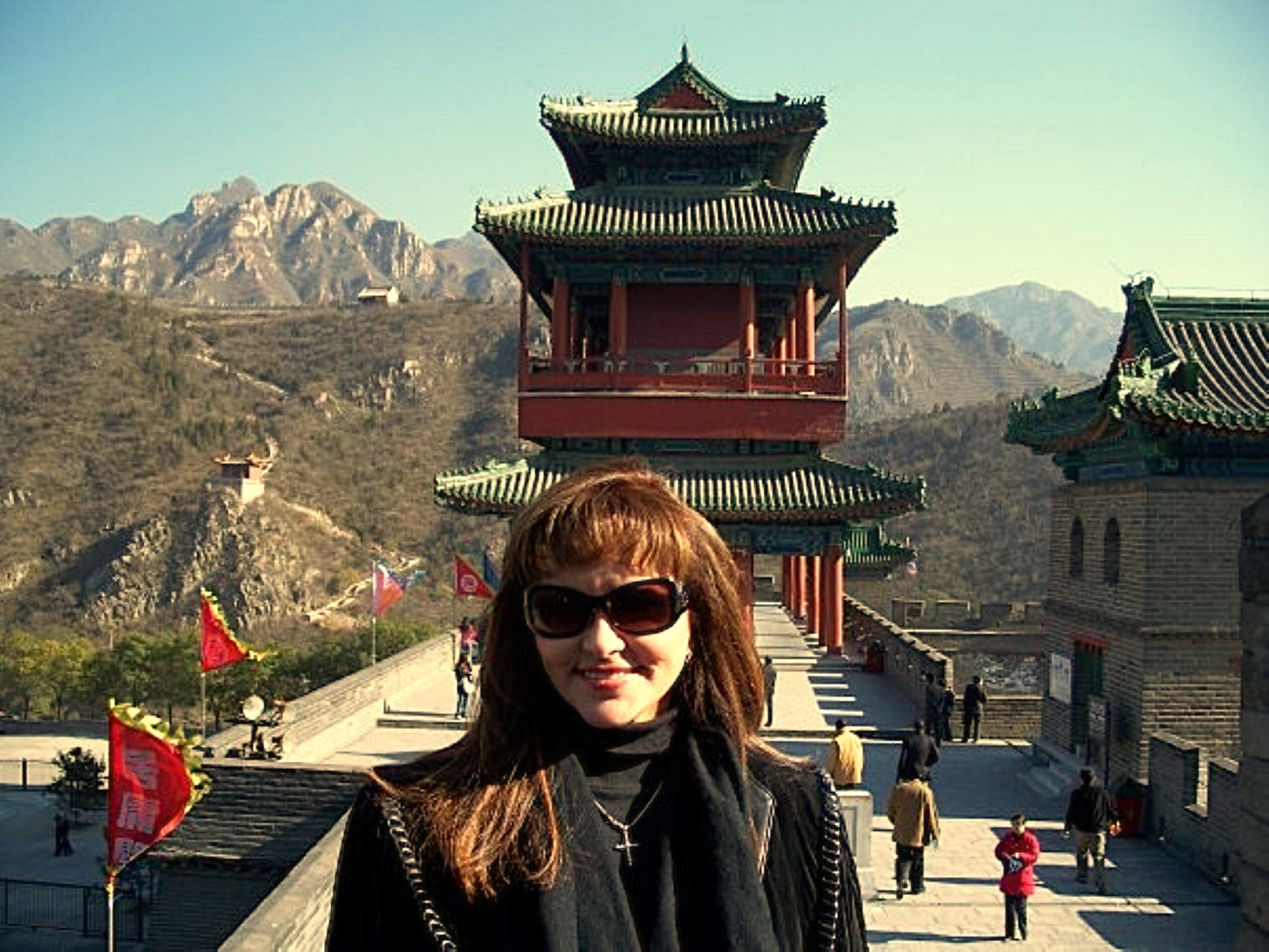 Woman with sunglasses and brown hair standing outdoors in front of traditional Asian pagoda-style buildings, mountains in background.