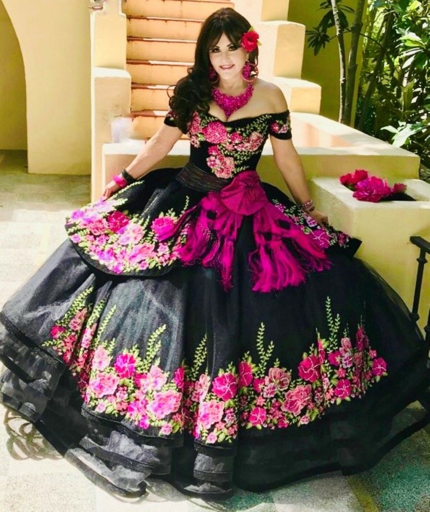 Woman in a black and pink floral dress sitting on a yellow staircase, surrounded by greenery, with flowers on her dress and nearby.