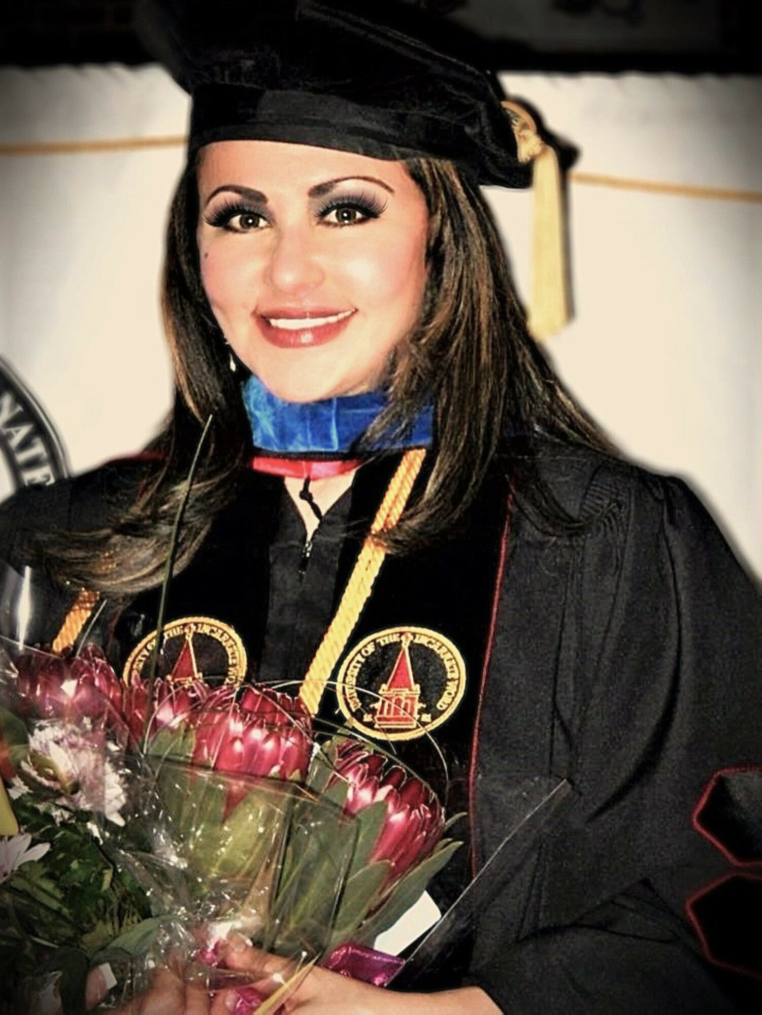 Woman in graduation gown and cap holding a bouquet of flowers, smiling, with a blurred background.