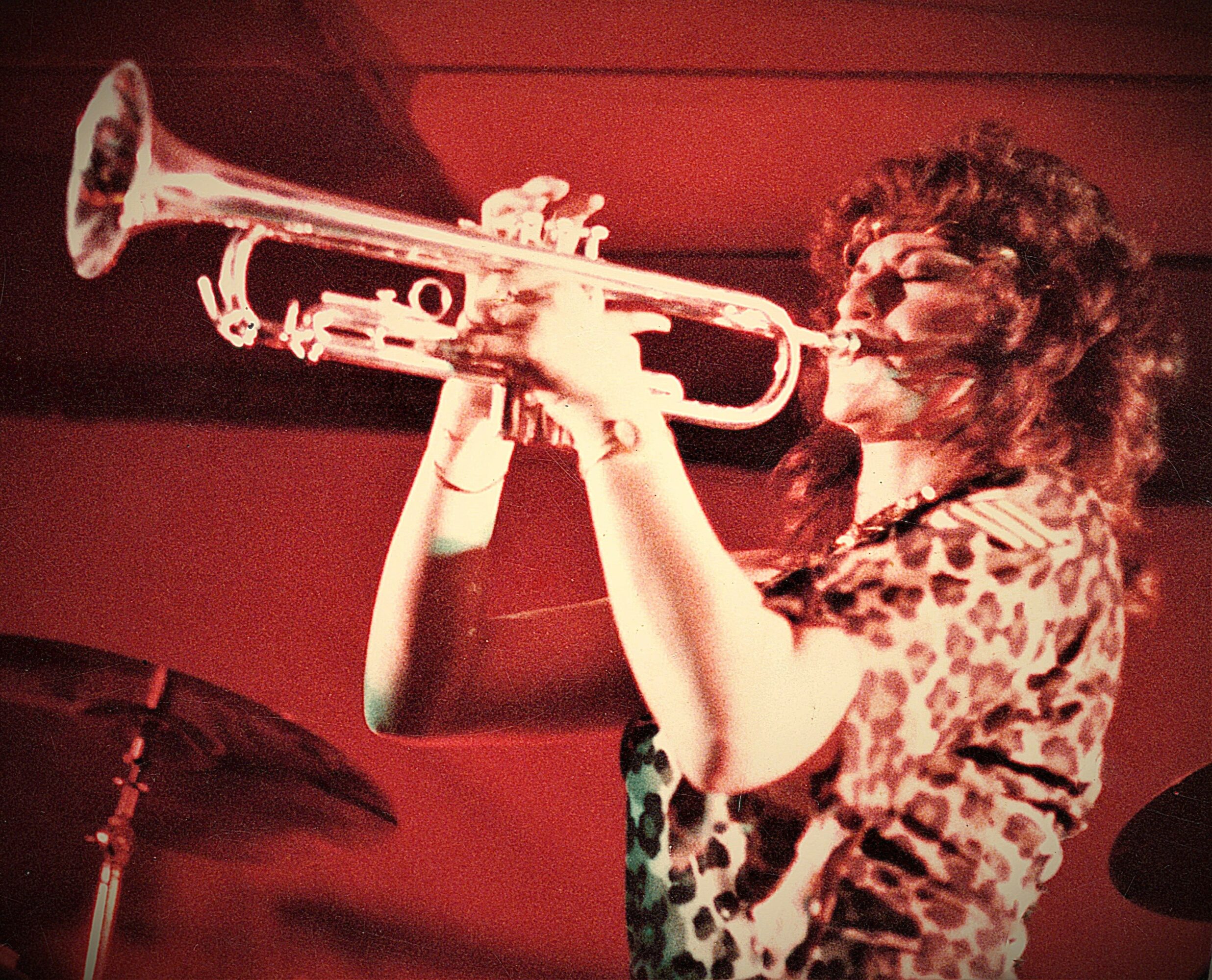 Person playing trumpet on stage with red background, curly hair, wearing patterned shirt.