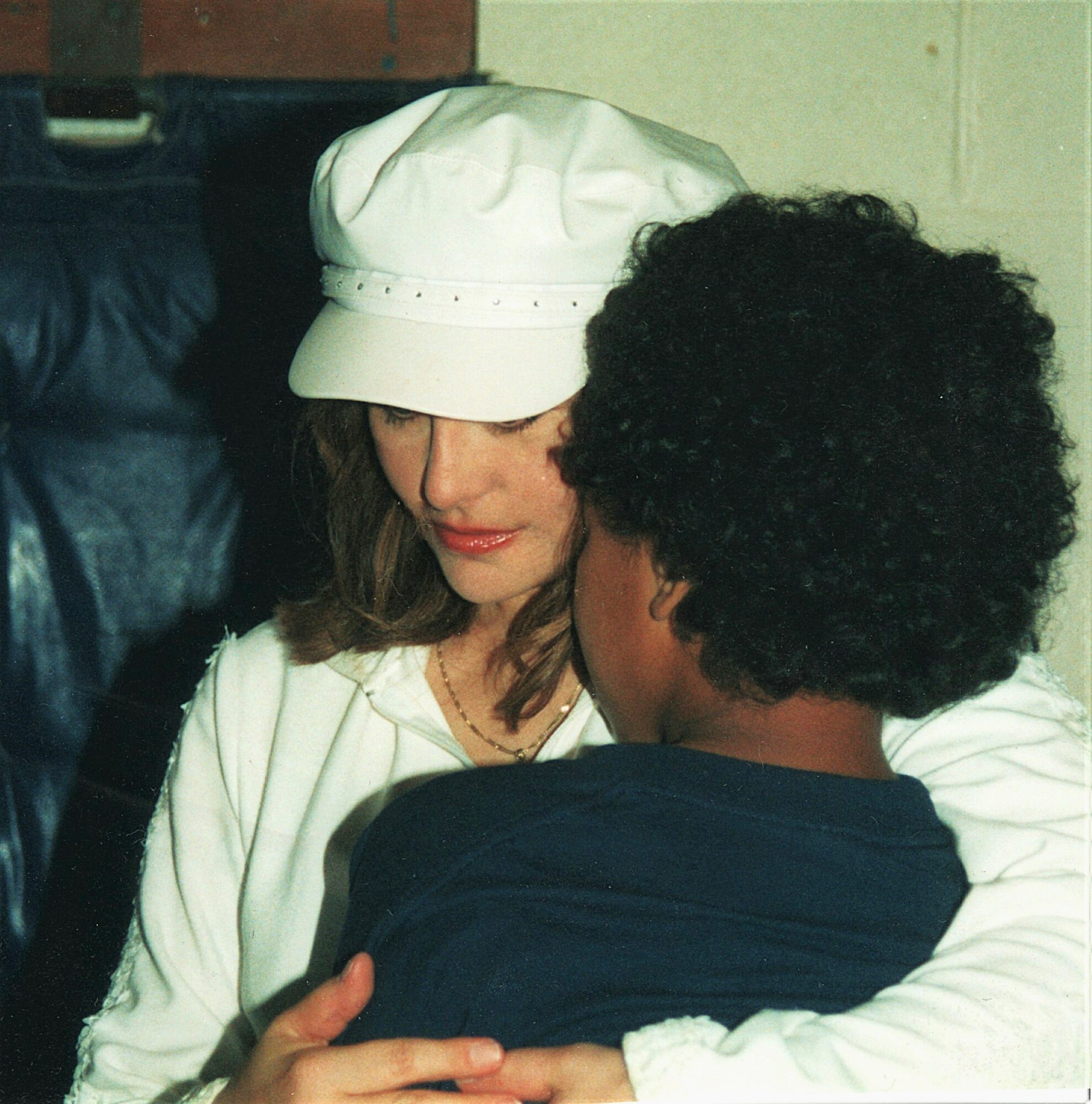 Woman wearing a white hat and white clothing hugging a person with curly hair in a dark shirt.