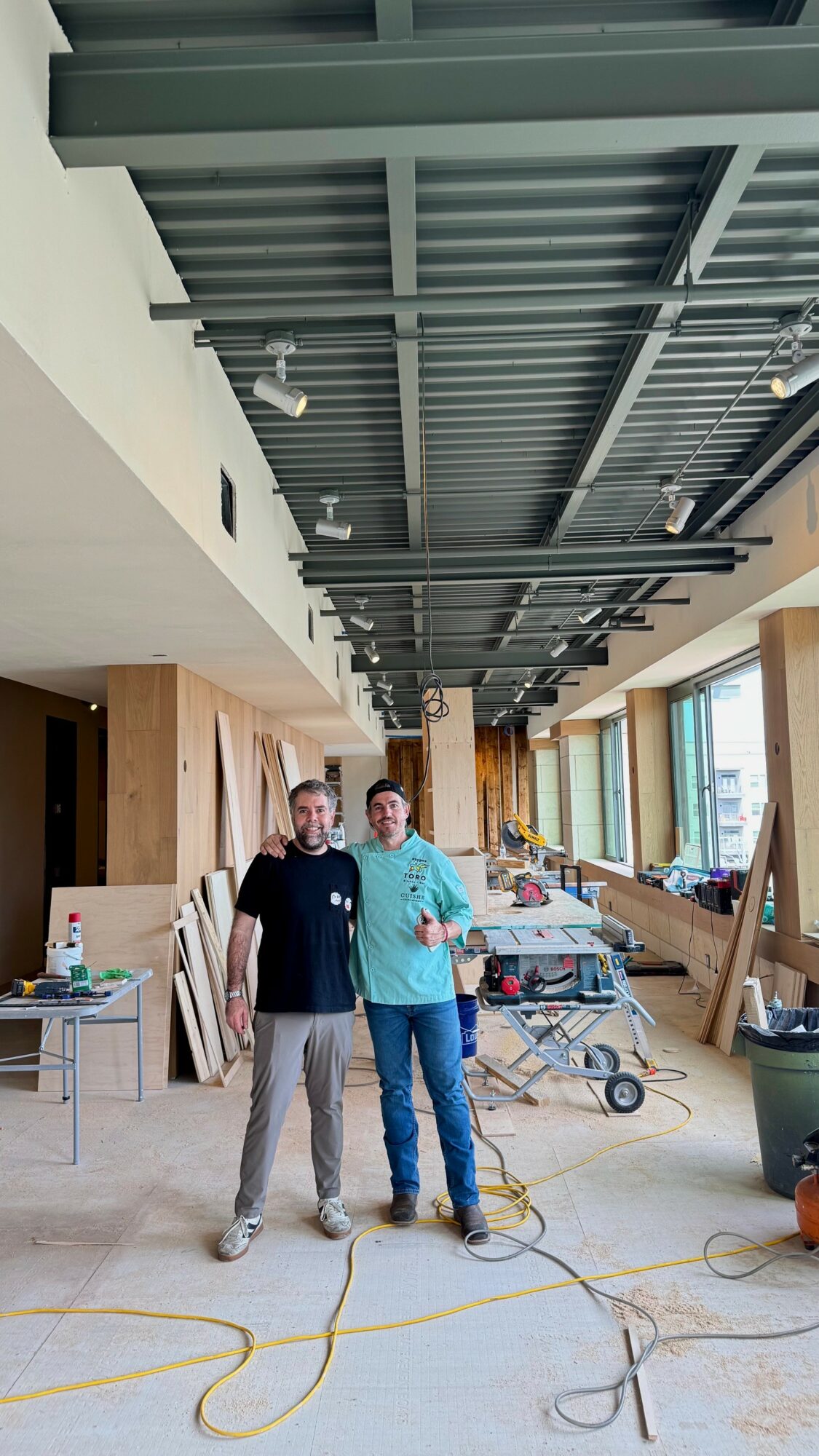 Two people standing in a spacious room with large windows, construction materials, and equipment visible, ceiling with exposed beams.
