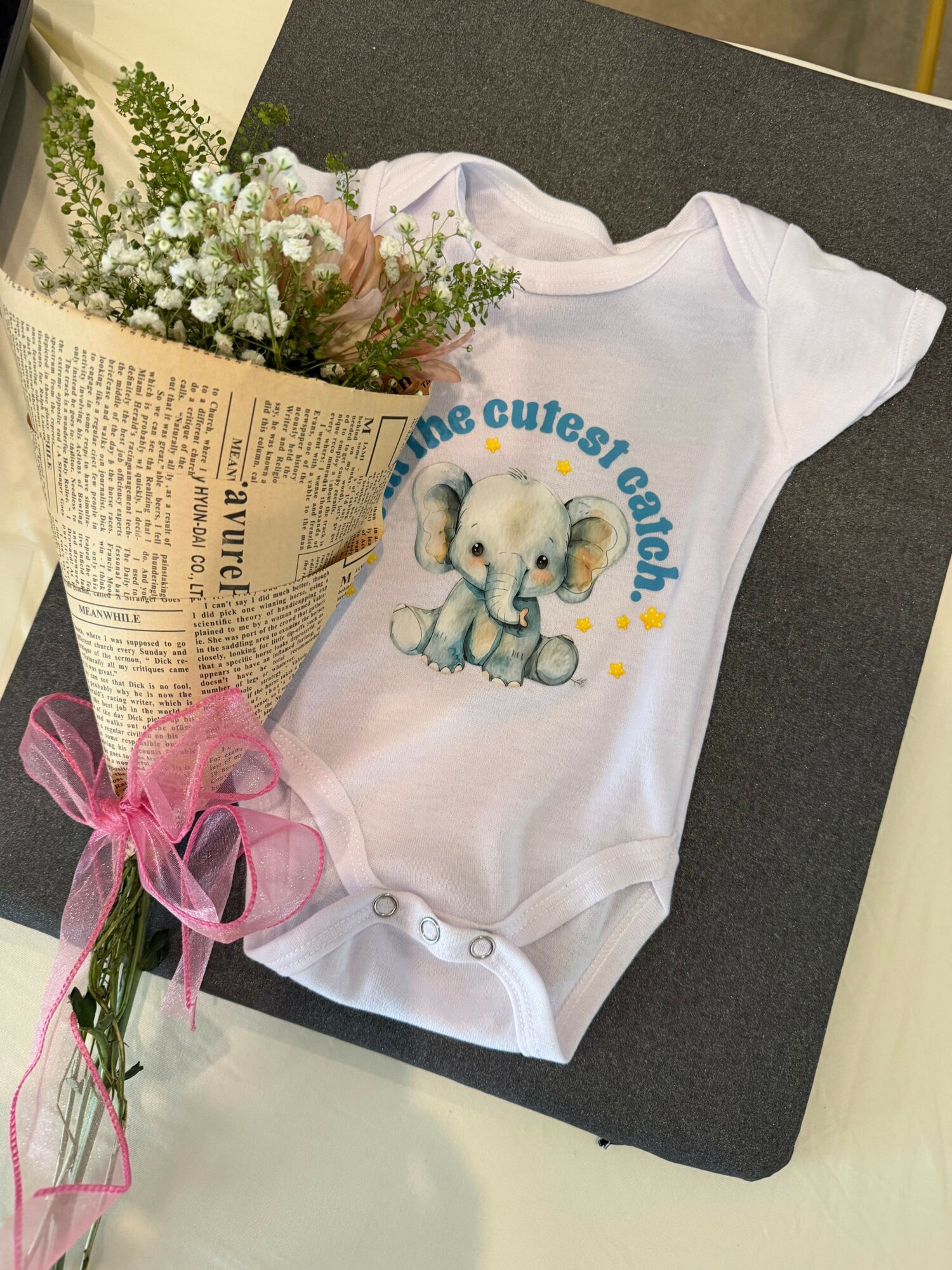 White baby onesie with a bear and text, pink ribbon, newspaper, and small white flowers on a dark surface.