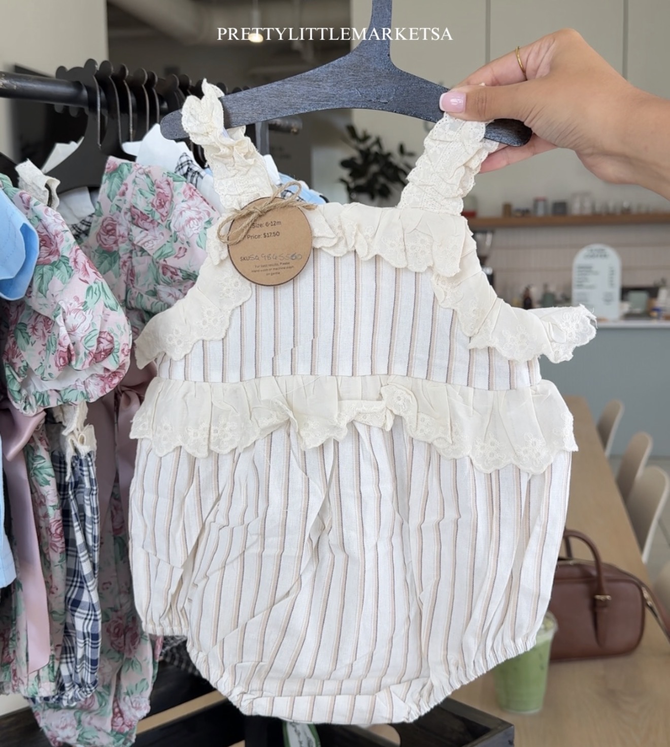 Person holding a cream-colored, striped baby romper with ruffles, hanging on a black hanger in a store. Background shows clothing and shelves.