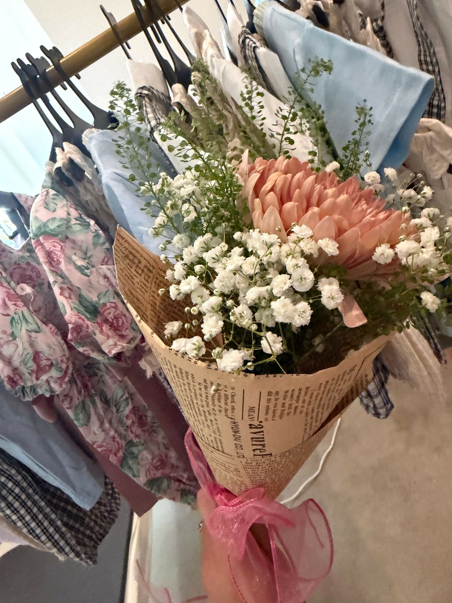 Bouquet of pink, white, and green flowers wrapped in newspaper with pink ribbon, placed on a floral cloth surface.