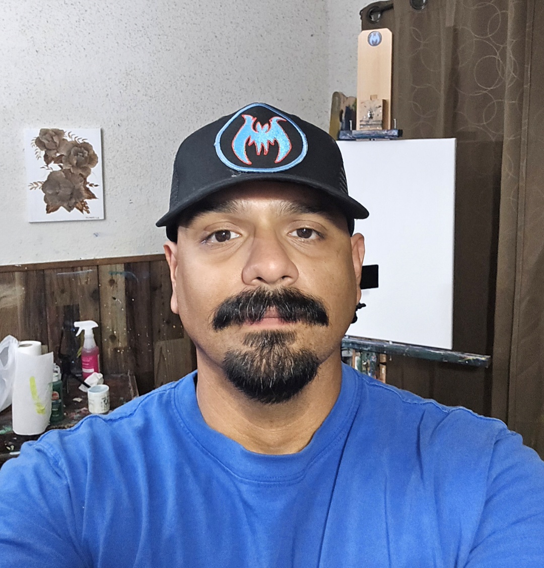 Man wearing a black cap with a blue and red logo, blue shirt, in a room with wooden walls and various objects