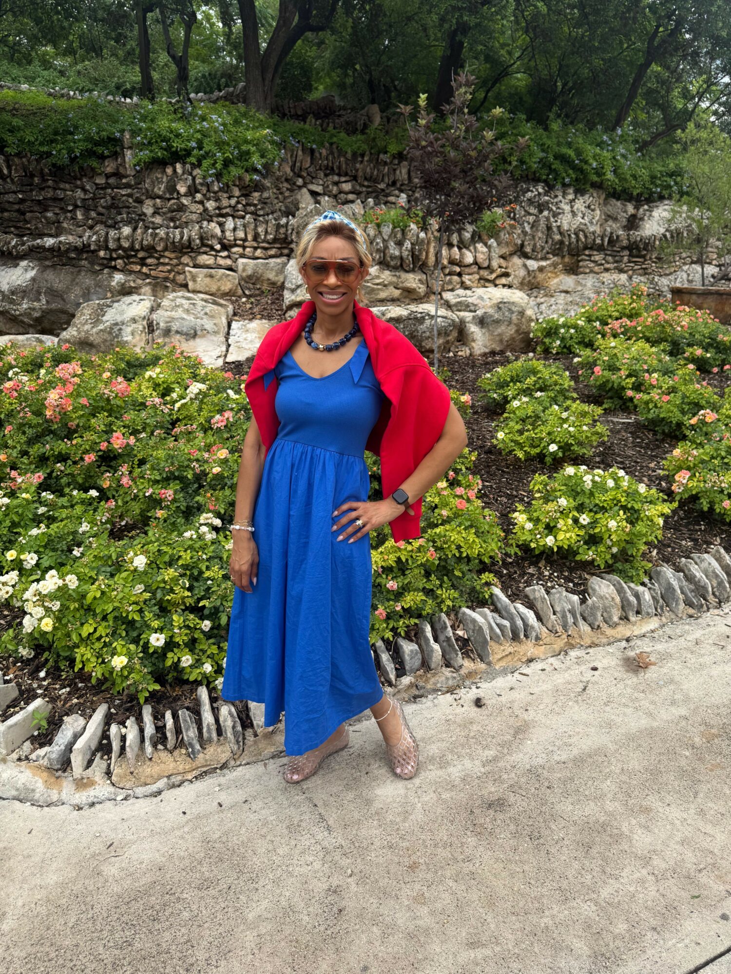 Woman in blue dress and red shawl standing outdoors with greenery and stone wall behind her.