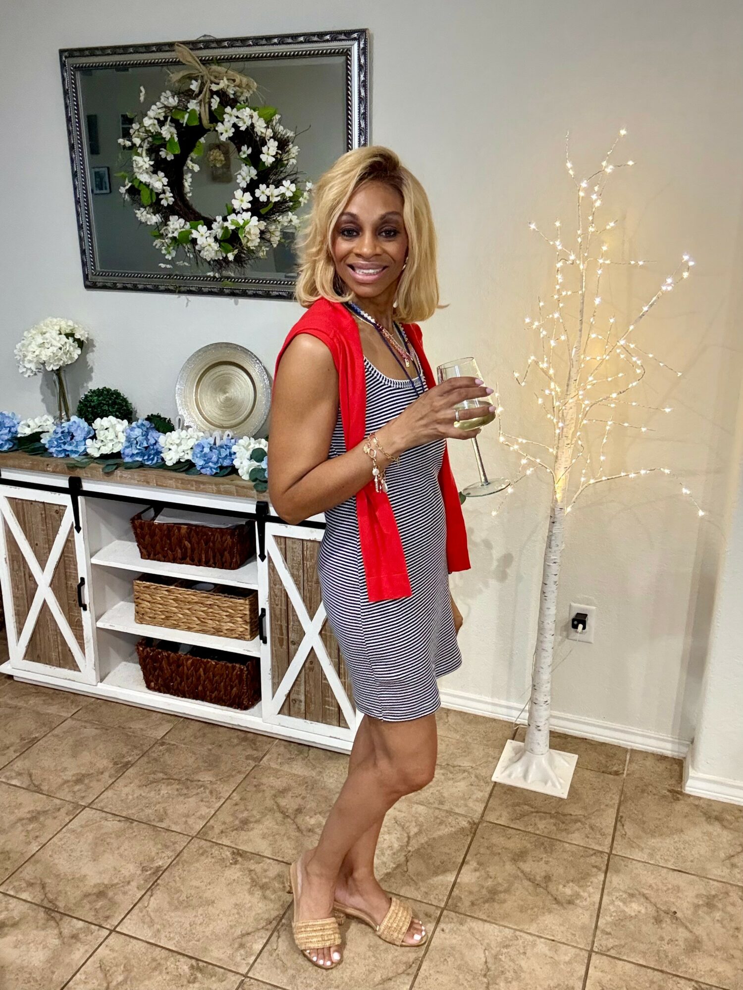 Woman in red scarf and gray dress holding a glass, smiling indoors with decorated shelf and illuminated tree in background.
