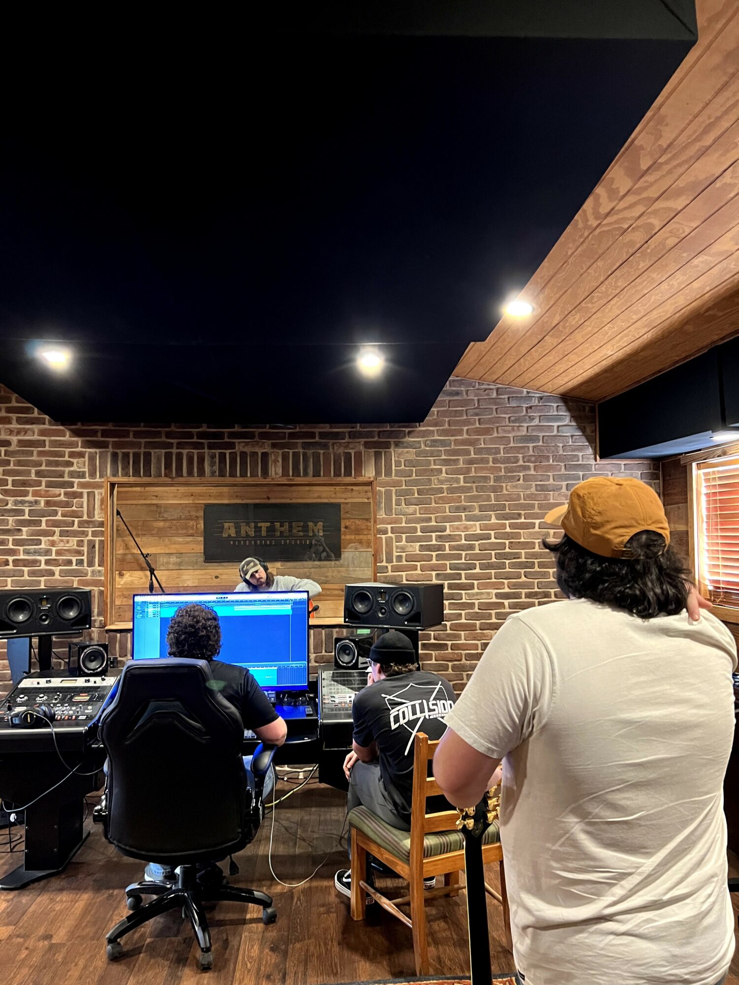 People in a recording studio with brick walls, a computer, and audio equipment, one person wearing a cap and white shirt.