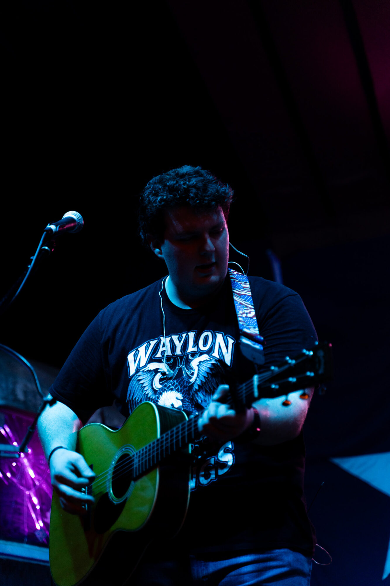 Person playing guitar on stage with microphone, dark background, blue and purple lighting, wearing a black t-shirt, curly hair
