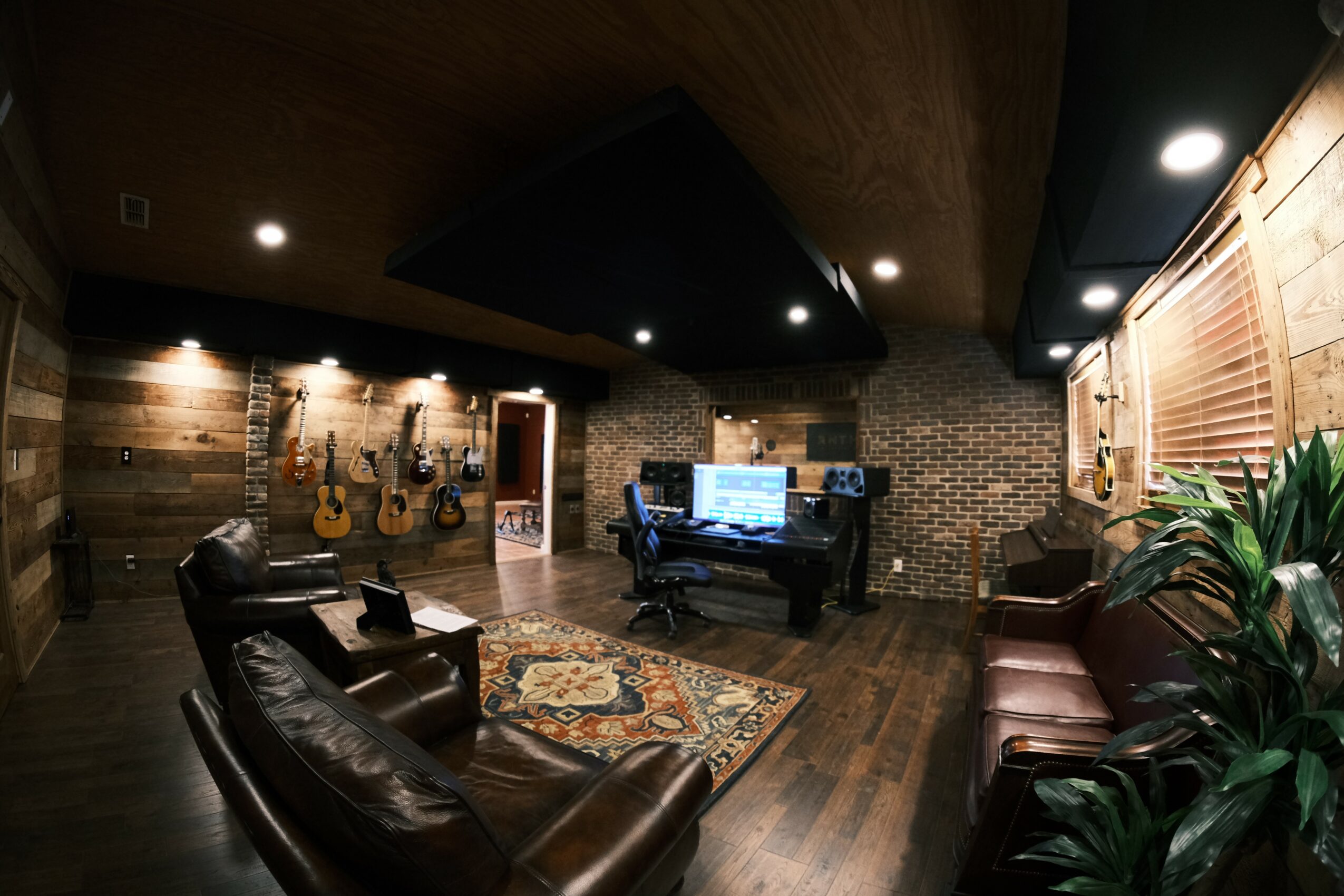 Music studio with guitars on wall, piano, leather chairs, and wooden walls, illuminated by ceiling lights.