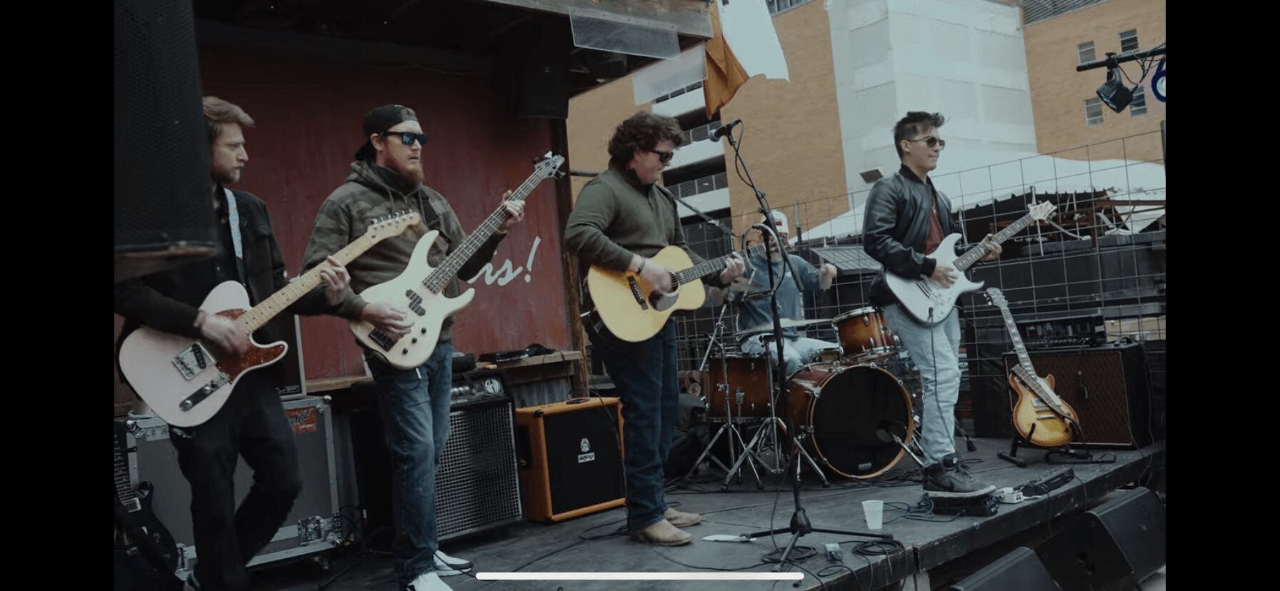 Group of five musicians performing on an outdoor stage with guitars and drums.