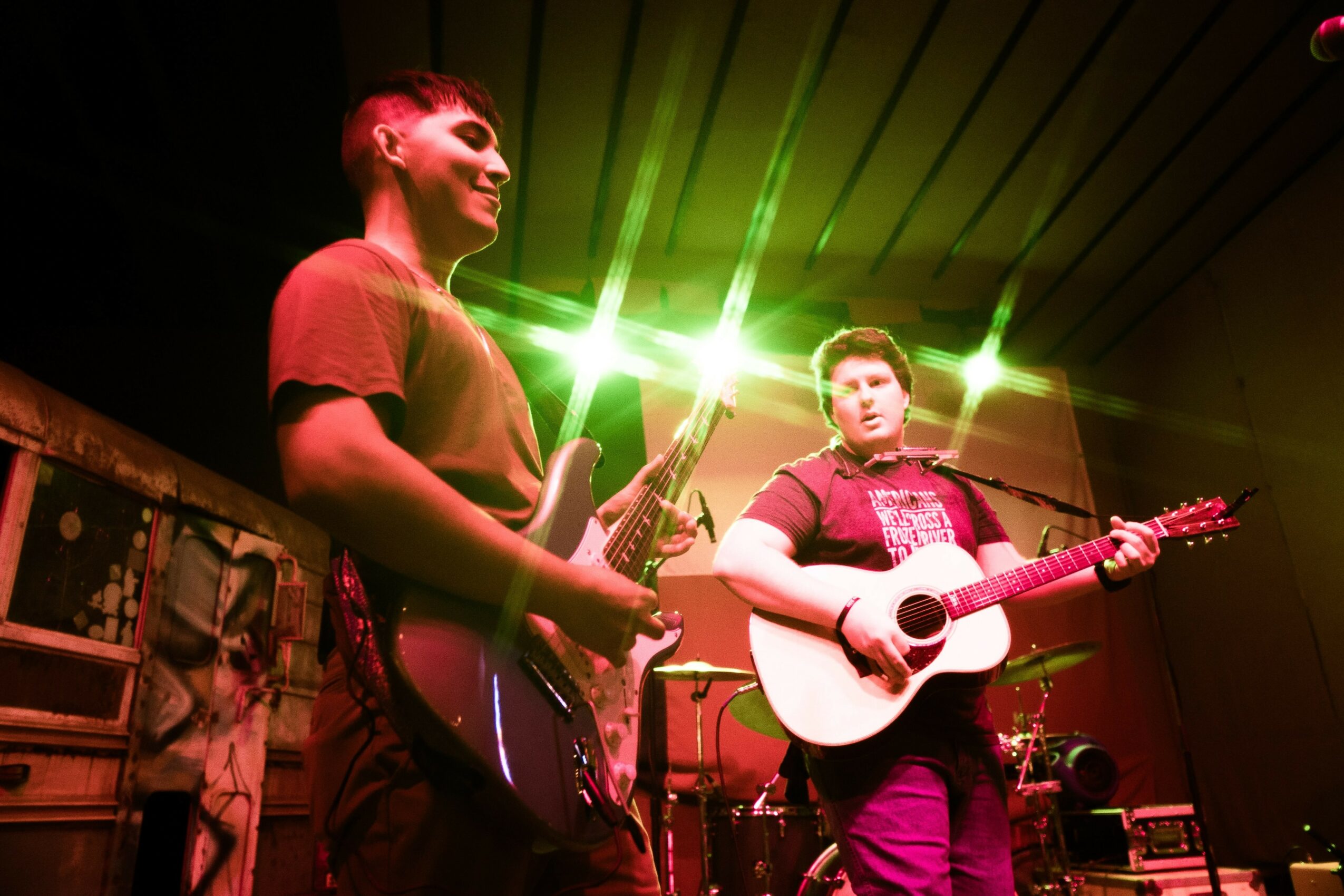 Two musicians perform on stage, one playing guitar and smiling, the other singing and playing guitar, with stage lights overhead.