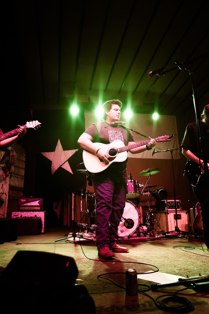 Musician playing guitar on stage with green lights and star decorations, surrounded by band equipment.