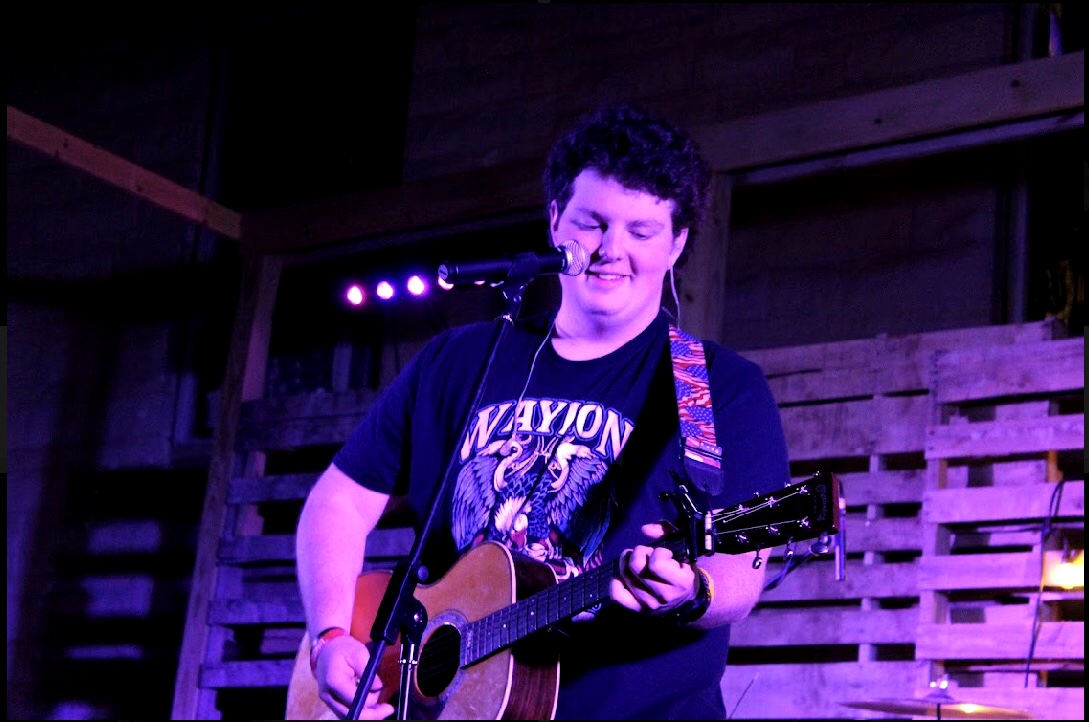 Person with curly hair singing into a microphone while playing an acoustic guitar on stage.