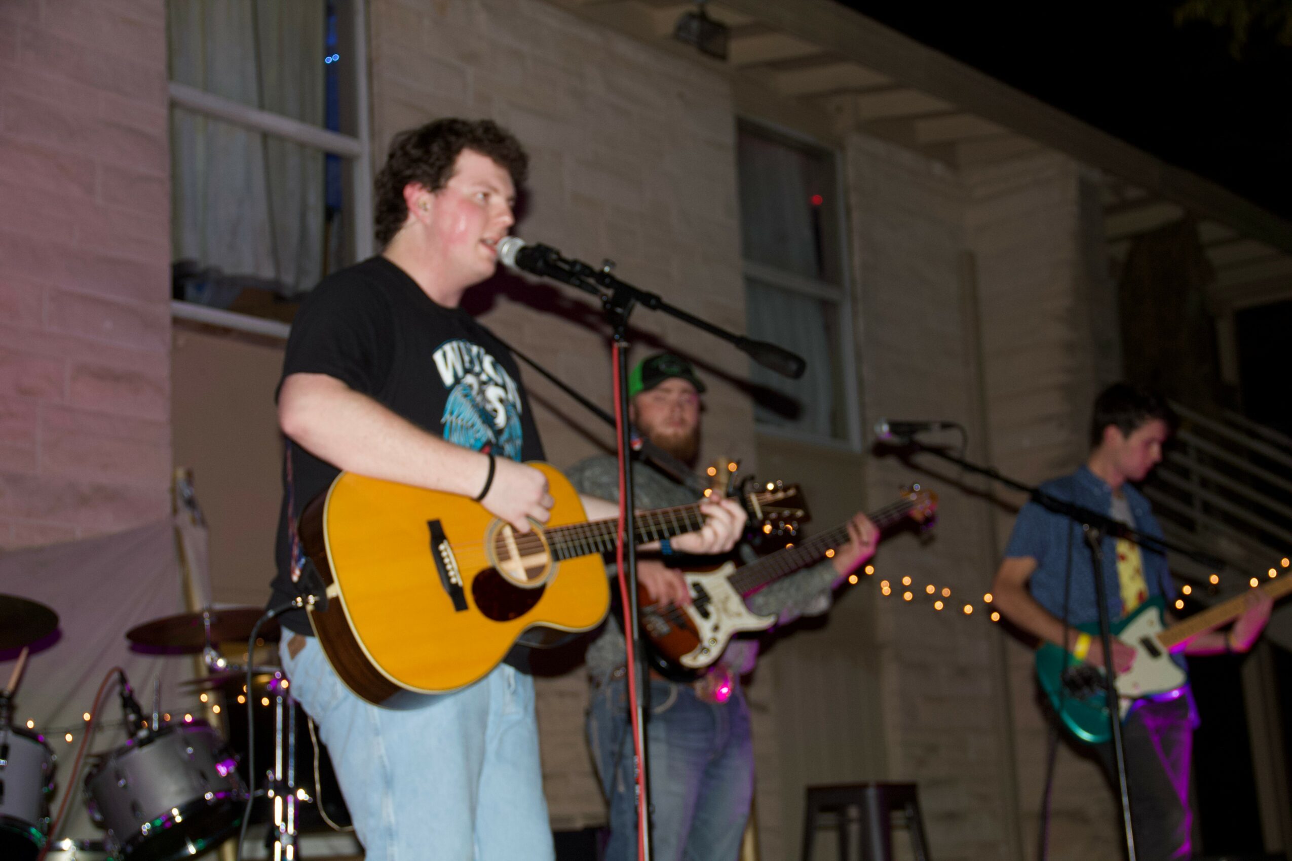 Three musicians perform on stage with guitars, microphone, and drums, in front of a building with string lights.