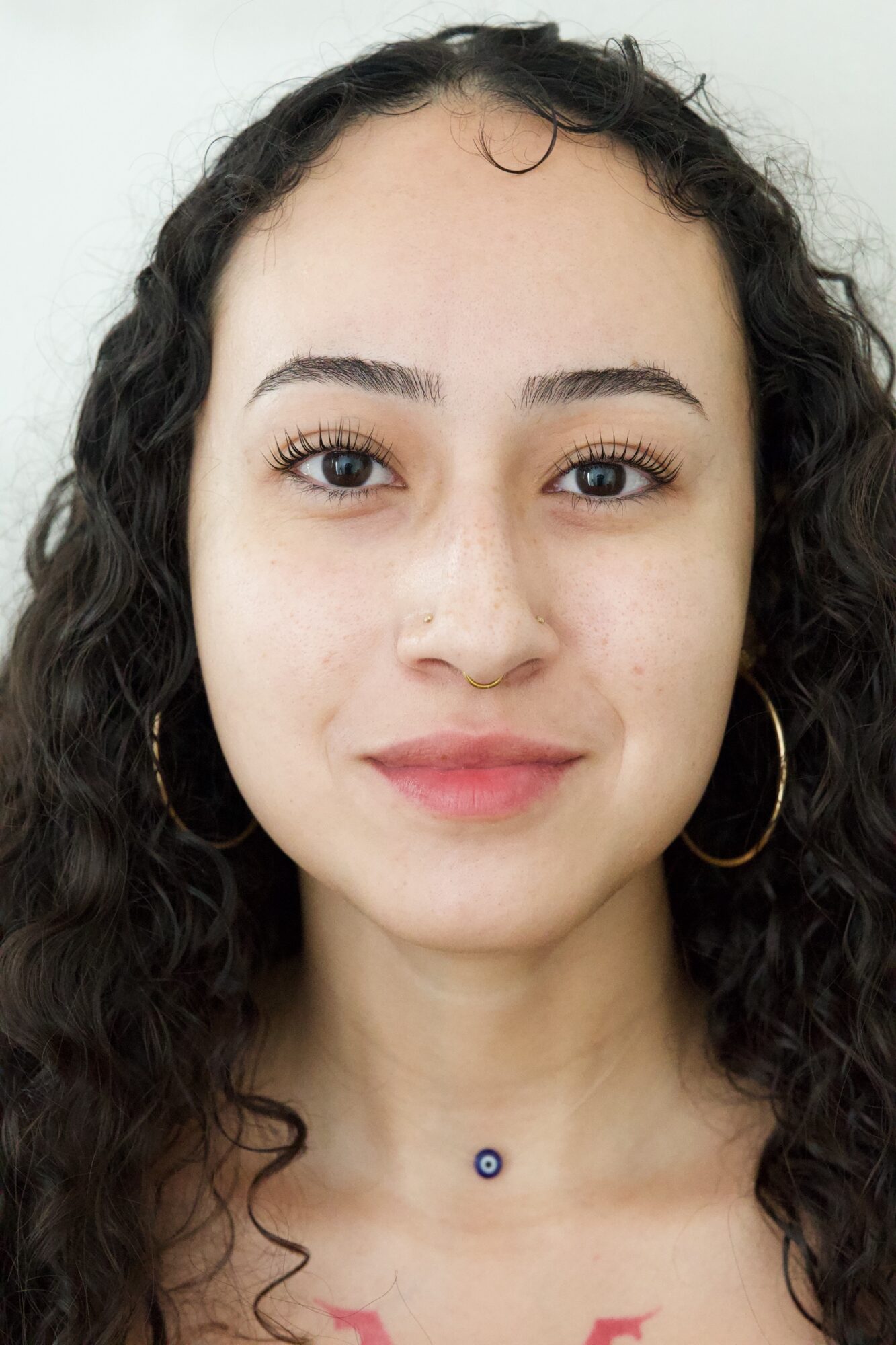 Young woman with curly hair, hoop earrings, and a small necklace, smiling softly against a plain background.