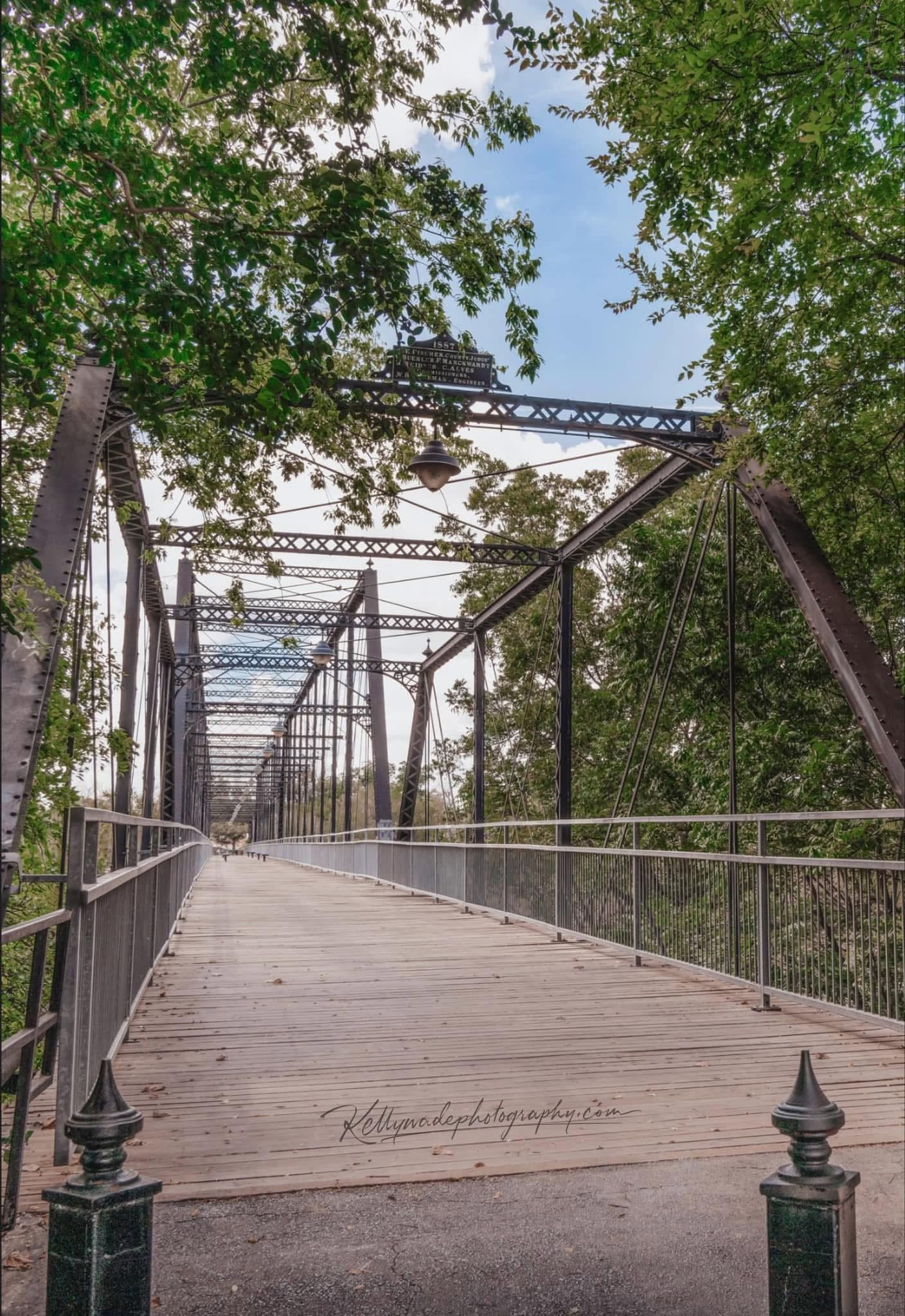 Wooden bridge with metal railings surrounded by green trees, sky visible above, and a pathway leading into the distance.