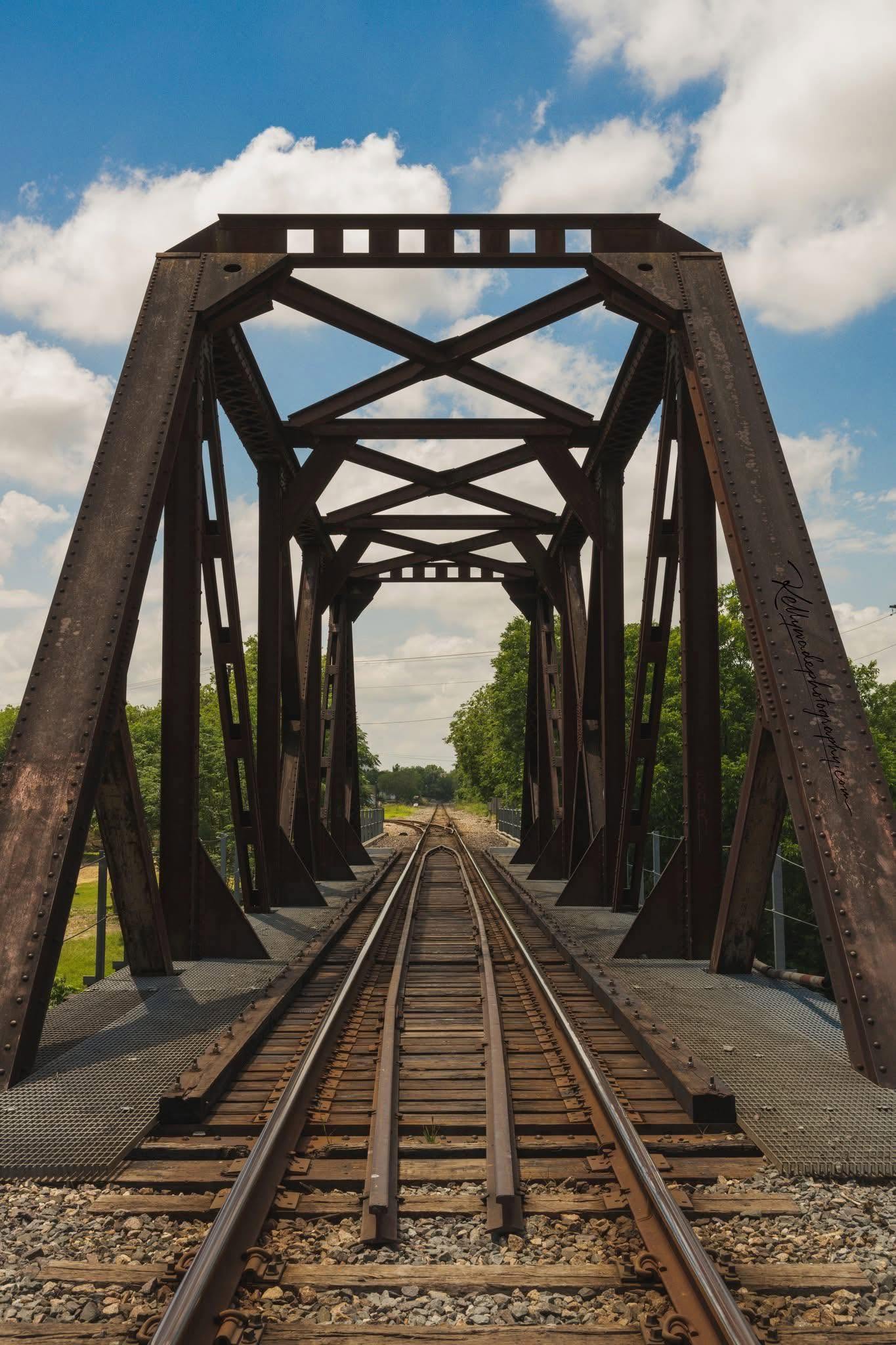 View of a railway bridge with metal framework over train tracks, under a partly cloudy sky.