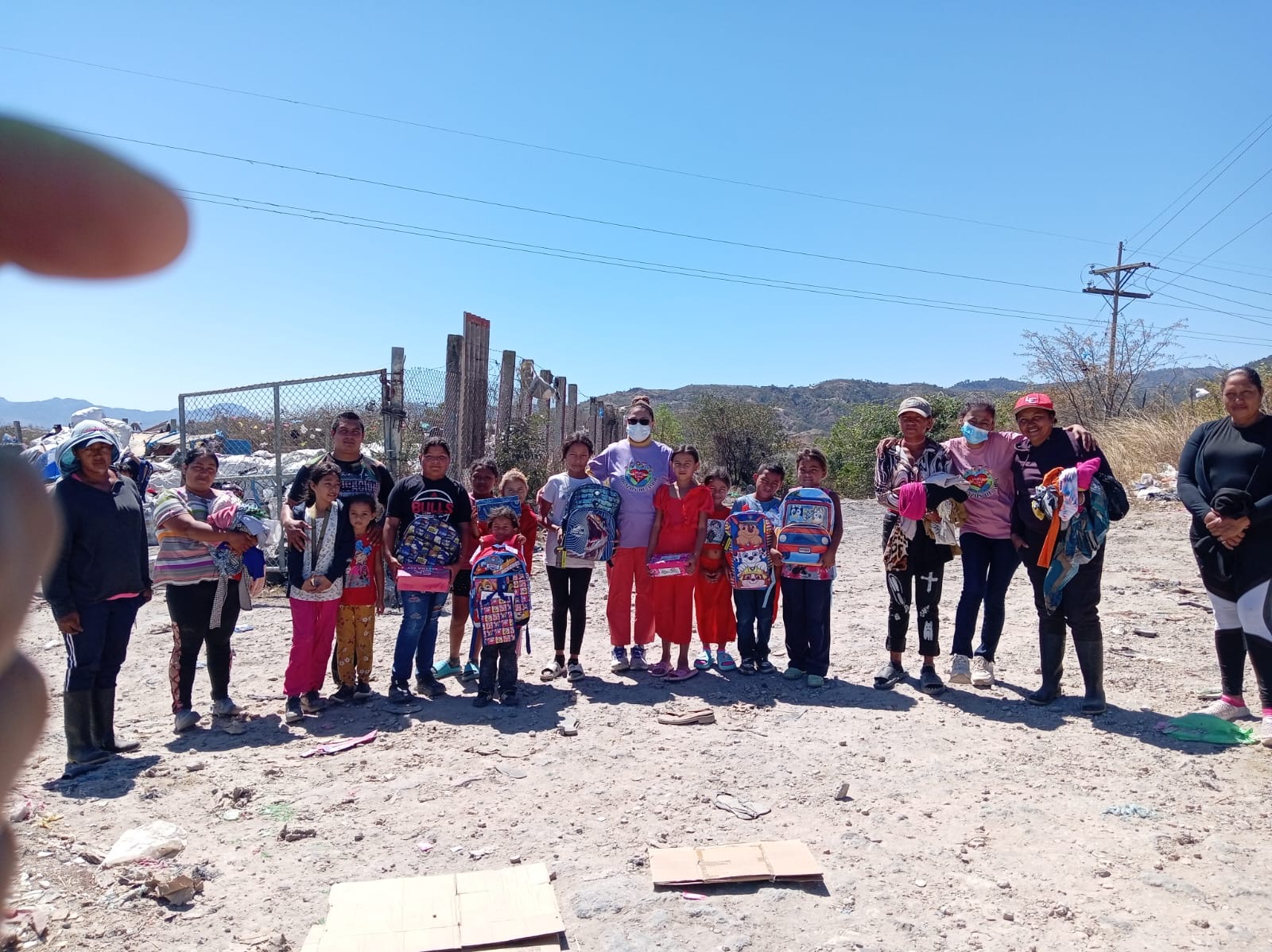 Group of children and adults outdoors on a sunny day, standing on a dirt ground with a fence and hills in background.