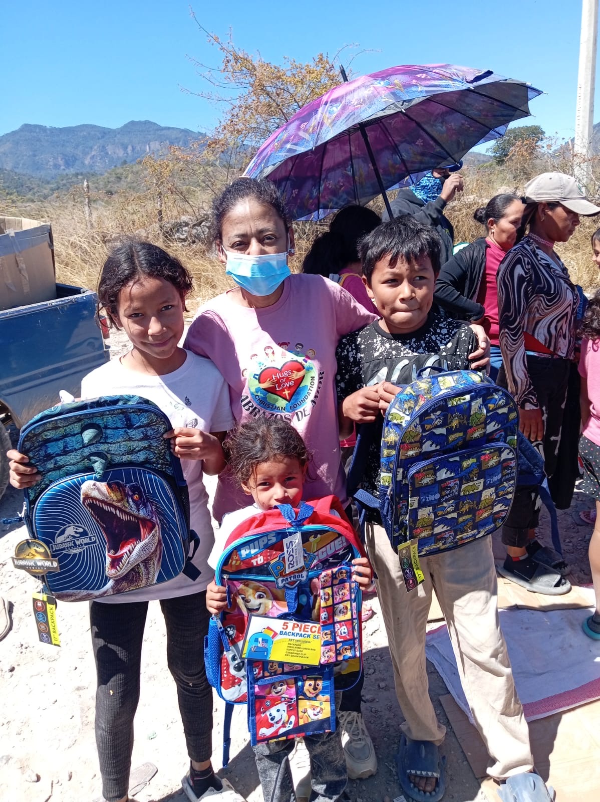 Group of children and a woman outdoors, some holding backpacks, with mountains and clear sky in background.