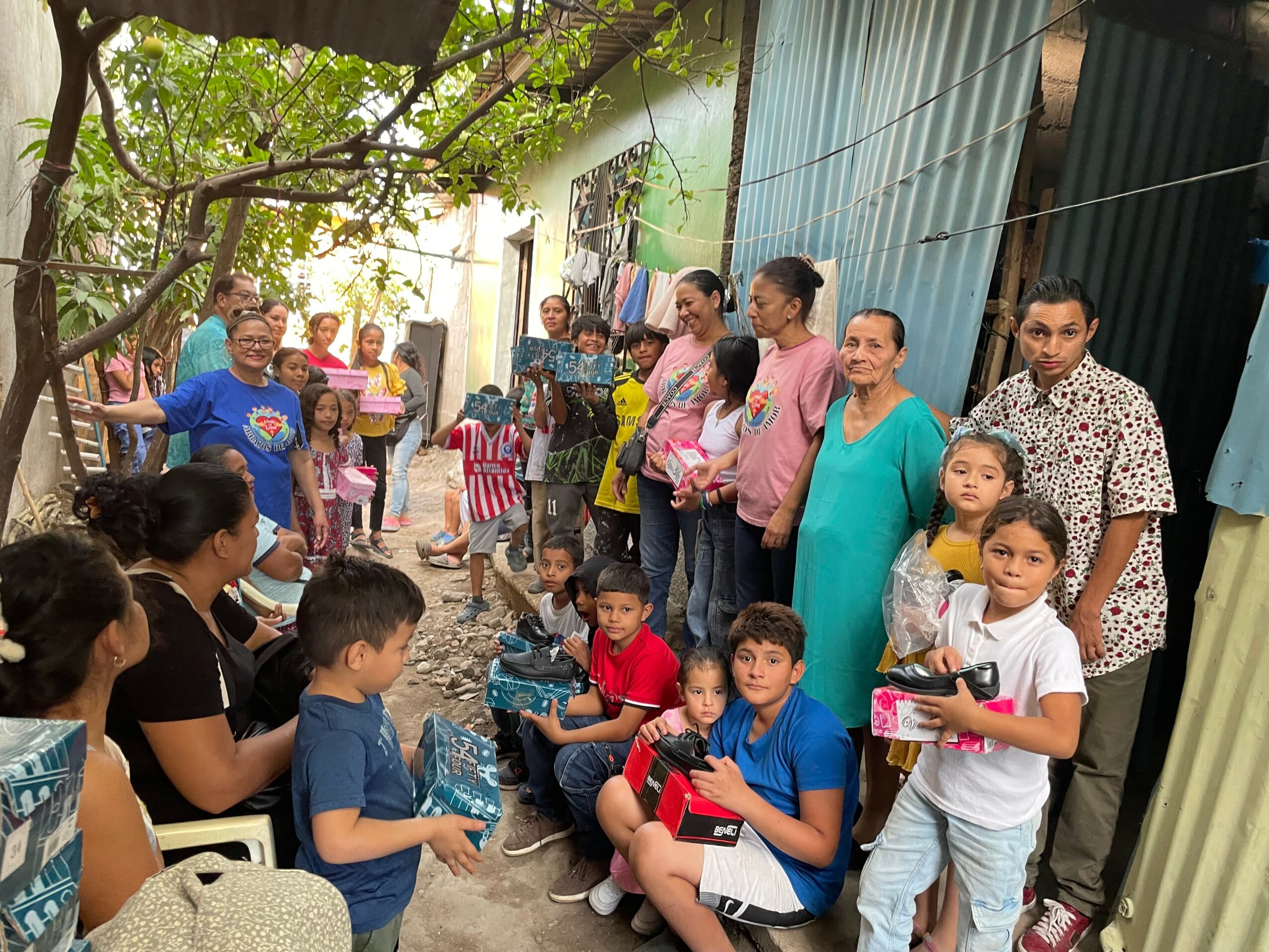 Group of children and adults gathered outdoors, some sitting and some standing, near a building and trees.