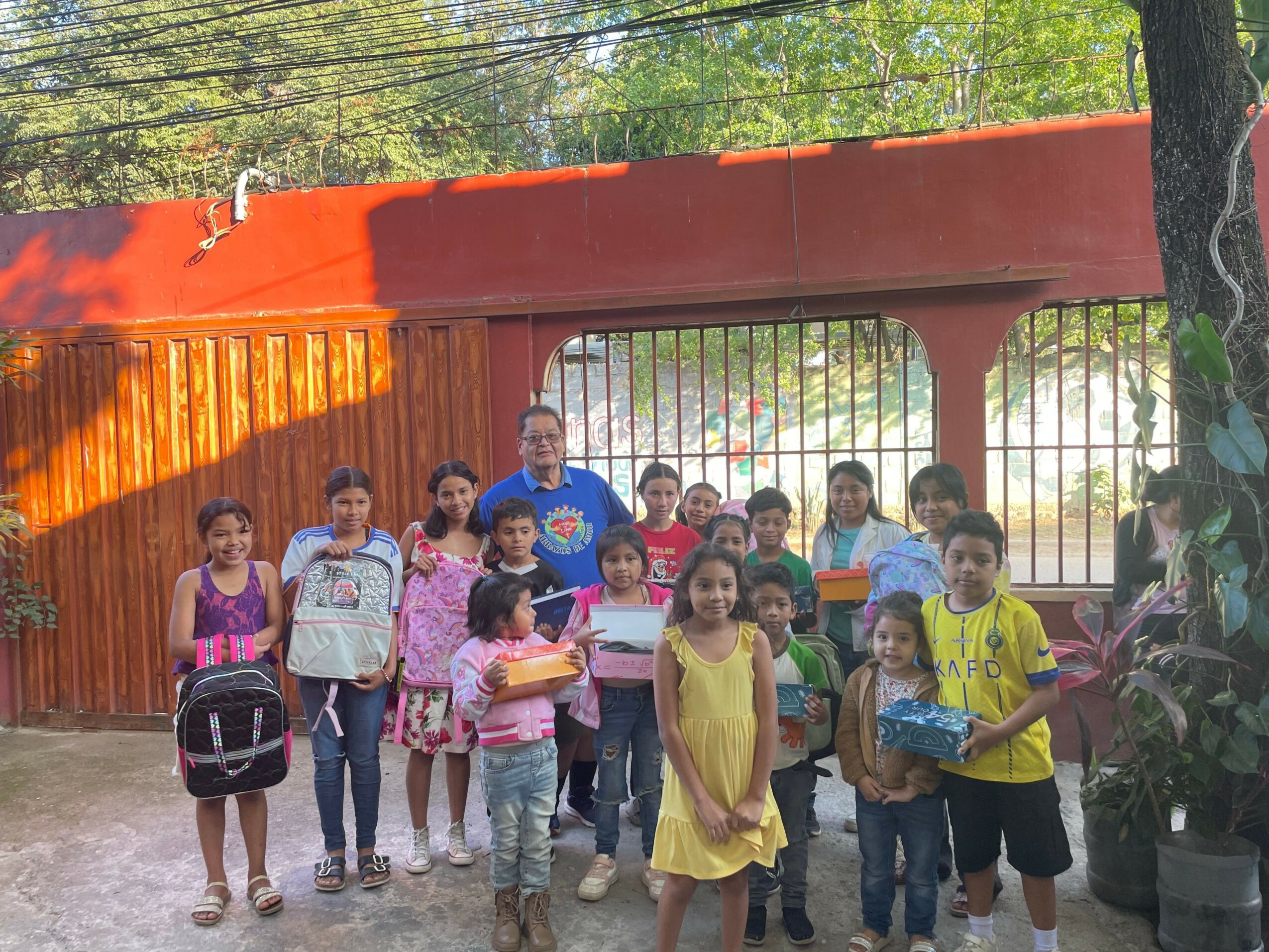 Group of children and an adult standing outdoors in front of a red wall and gate, some holding backpacks and bags.