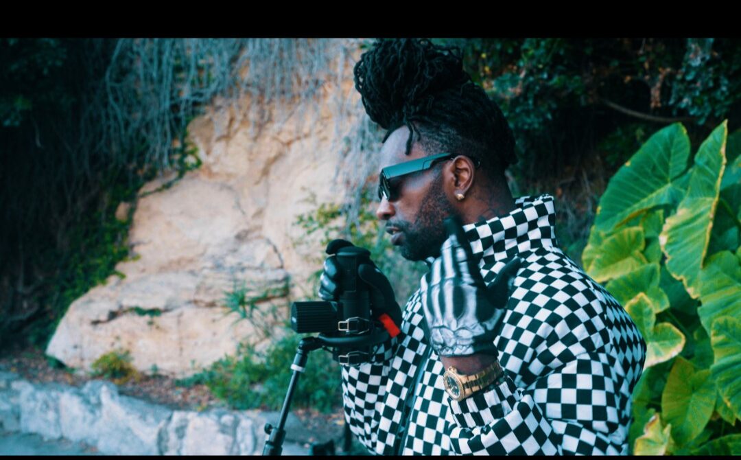 Person with sunglasses and checkered jacket holding a camera outdoors near greenery and rocks.