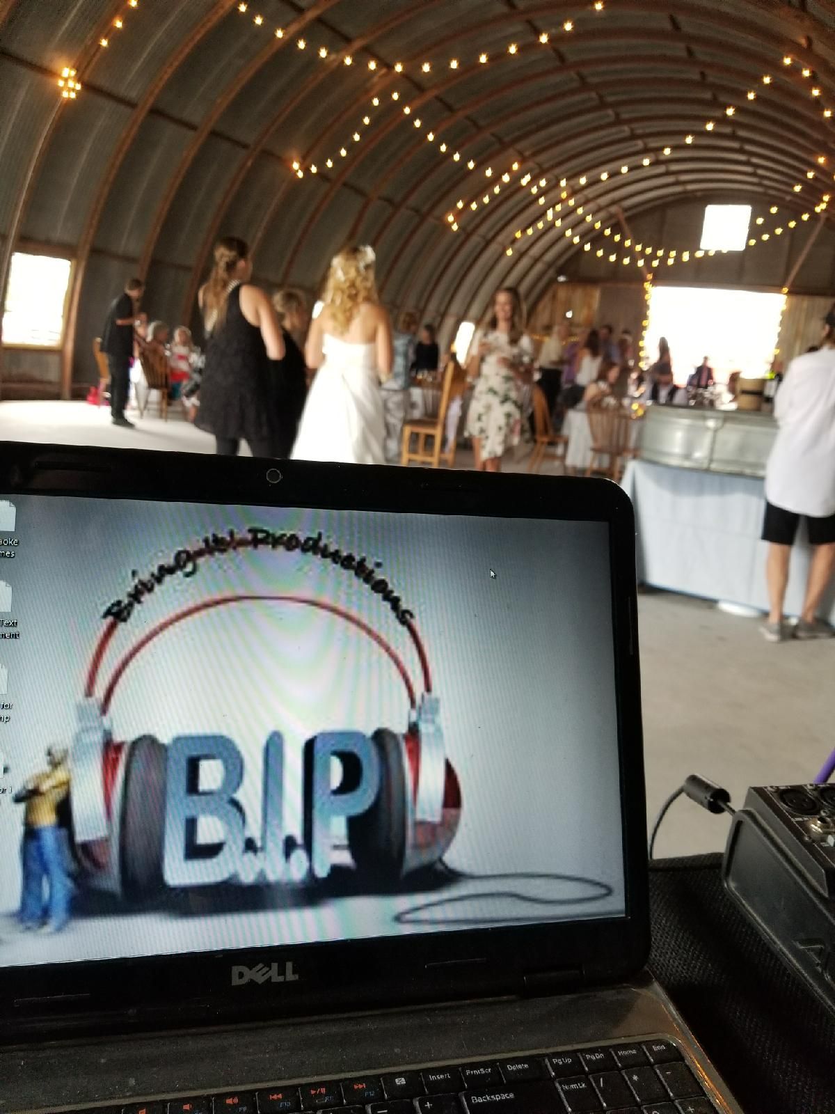 People dancing and socializing in a large, decorated indoor space with string lights, seen from behind a laptop displaying a logo.